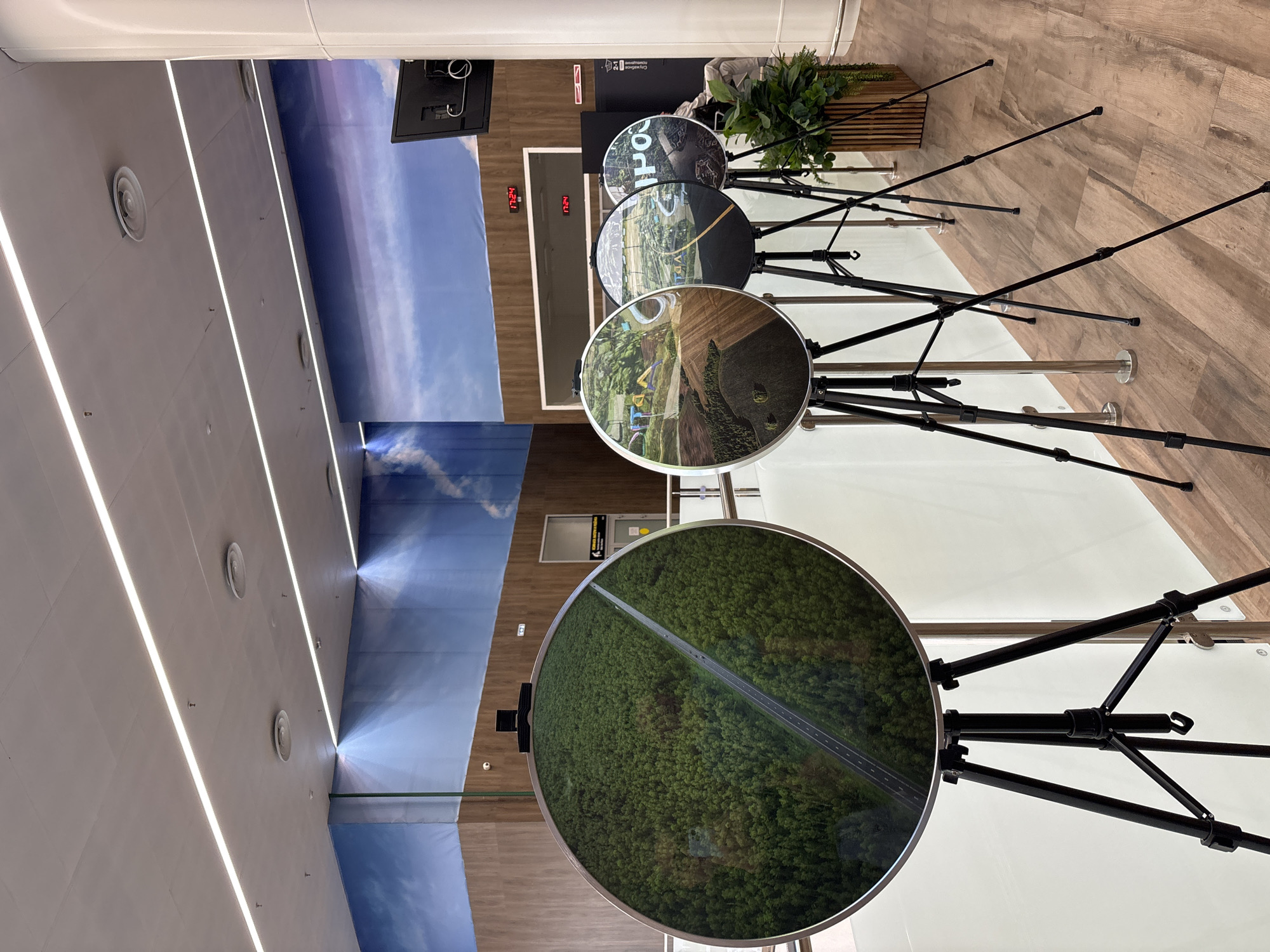 This media file shows a modern interior space, possibly an exhibition hall or museum, with a sky-themed mural on the wall. Several circular displays, mounted on black stands, are arranged in a row. Each display features a photographic image, likely aerial views, of diverse landscapes. The top display shows text that includes "PO", suggesting a brand or exhibit name. Below this, another display shows a landscape with a road and greenery, possibly indicating a rural or suburban setting. The third display from the top is a close-up of a vibrant green forest with a road running through it. The lighting in the room appears to be artificial, with long, bright fluorescent lights running along the ceiling and spotlights illuminating the mural. The floor is a light-colored wood laminate. The overall ambiance is clean, contemporary, and designed for visual presentation. There are no people visible in the image, and the activity appears to be static, focused on the displays. Two red digital clocks display "17.24" on the wall.