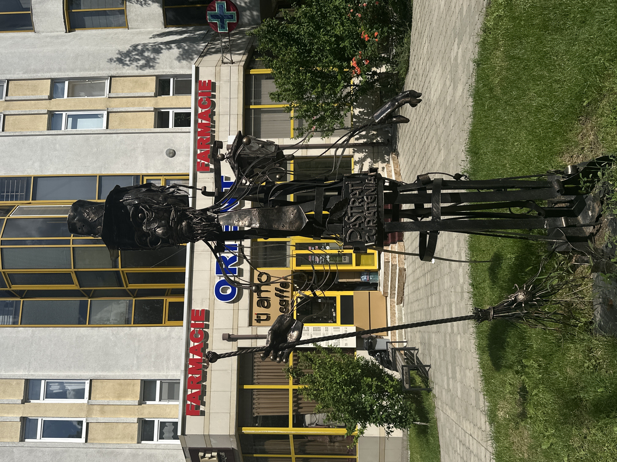 A metal sculpture of a witch or sorcerer stands in front of a building with the signs "FARMACIE" in red letters. The sculpture is made of dark metal and depicts a figure wearing a top hat and holding a broom. The building has large yellow-trimmed windows and appears to be a commercial establishment. The ground in front of the building is paved with grey interlocking stones, and there is a patch of green grass to the left of the sculpture. The sculpture is located in Chișinău, Moldova, specifically on Alexandru cel Bun Street.