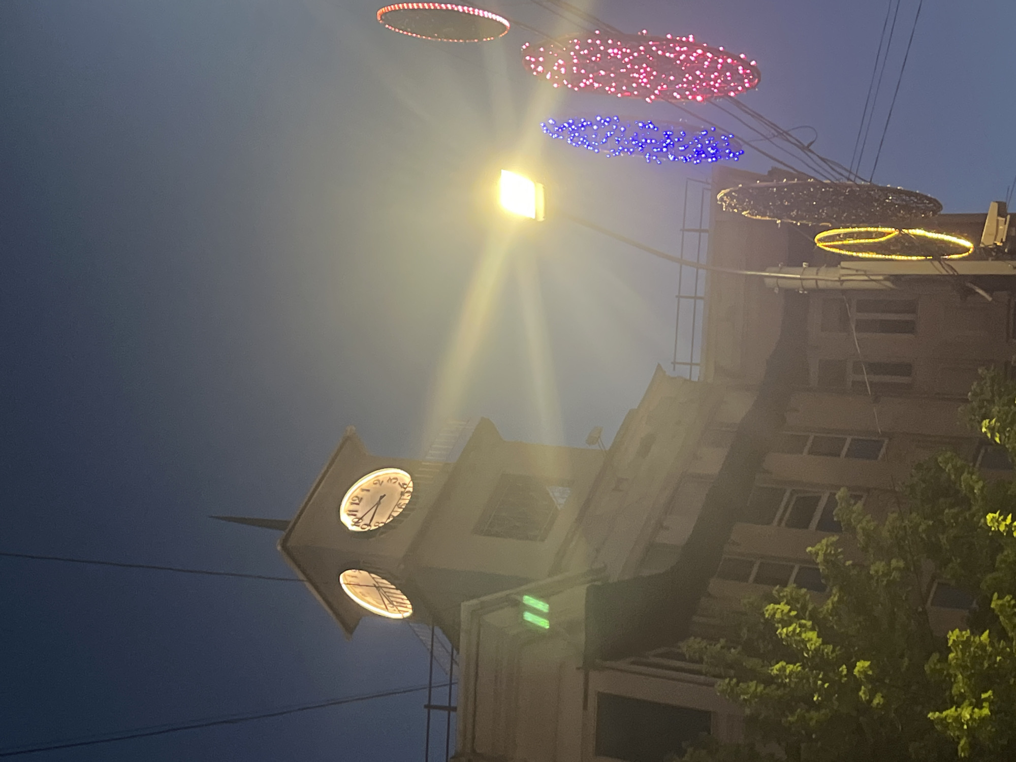 The image depicts a city street scene at dusk, with a clear night sky overhead. In the foreground, a clock tower is prominently featured, its illuminated clock faces displaying the time. The tower is angled, giving a sense of depth to the composition. Above the tower, decorative lights are strung across the sky, appearing as illuminated rings and chains of colorful lights in red and blue. A bright, square light source, possibly a street lamp or a projector, casts a strong beam of light downwards, creating a lens flare effect.
To the right of the clock tower, a multi-story building is visible, with windows reflecting the ambient light. A lush green tree with illuminated foliage is positioned in the lower right corner, adding a natural element to the urban setting. Overhead power lines are also visible, crisscrossing the sky.
The overall mood is festive and illuminated, suggesting an evening event or a decorated urban environment. The time of day is clearly evening or night due to the dark sky and the presence of artificial lighting. There is no visible text in the image. The setting appears to be an urban area, likely a public square or street, given the presence of a clock tower, buildings, and street decorations. The image is captured from a slightly low angle, looking upwards towards the sky and the illuminated structures.