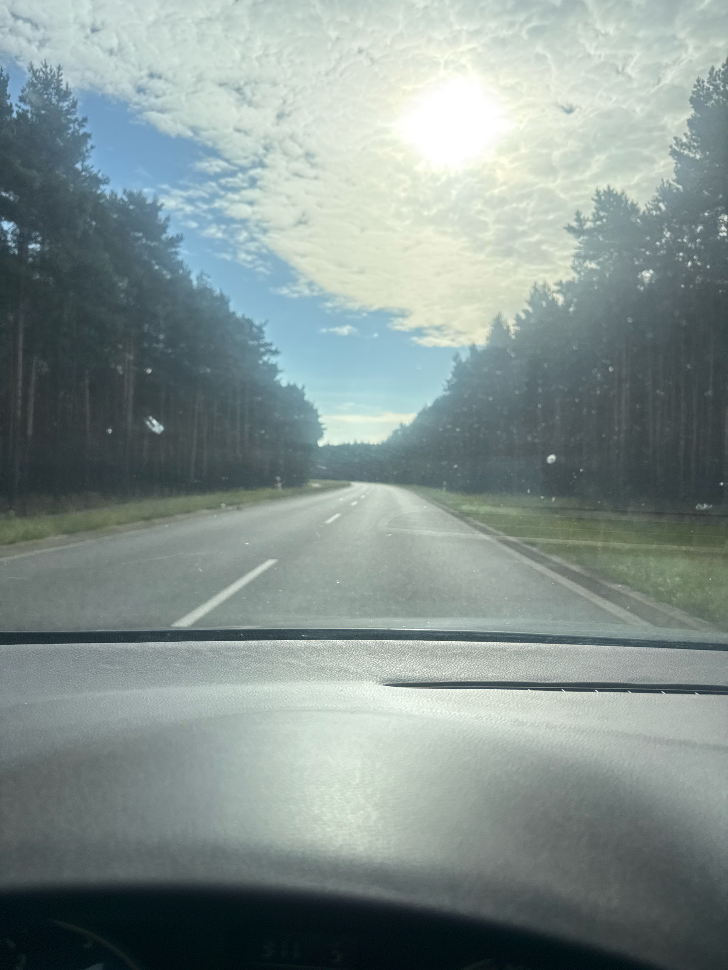 This media file is a photograph taken from inside a vehicle, looking forward through the windshield. The scene is a paved road winding through a dense forest of tall, dark green pine trees on either side. The road has a solid white line marking its edge on the right and a dashed white line on the left, indicating a two-lane road. The sun is shining brightly in a partly cloudy sky, with scattered white clouds that appear somewhat textured. The sun is high in the sky, suggesting it is daytime, likely late morning or early afternoon. There are some small, circular spots visible on the windshield, which are likely water droplets or dust. The perspective suggests the vehicle is in motion, traveling along the road. The overall mood is serene and natural, with the sunlight filtering through the clouds and trees creating a bright, clear atmosphere. The location is identified as Rudno, Poland. No people or specific objects other than the road and trees are clearly discernible. There is no visible text in the image.
