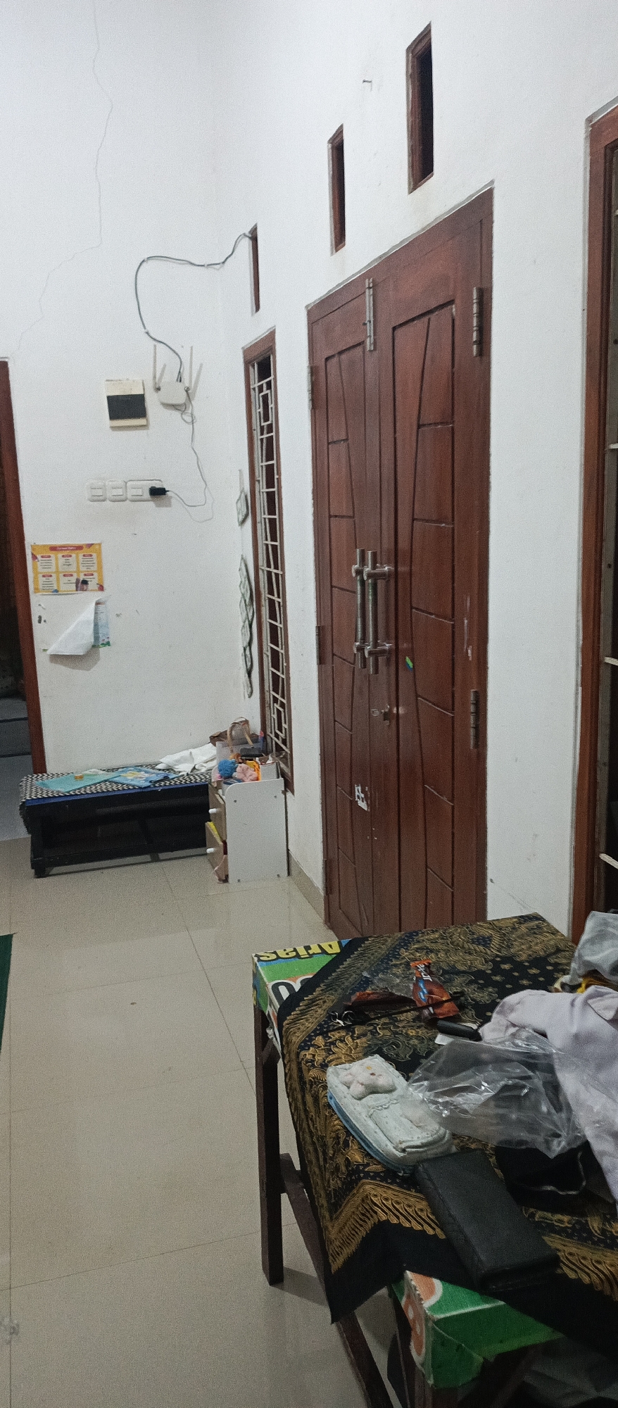 The image depicts an interior scene, likely a residential hallway or room, in Arjasa, Indonesia. A large wooden double door with vertical paneling and metallic handles dominates the right side of the frame. To the left of the door, a narrow, tall window with a decorative grille is visible. Further left, a white wall features electrical outlets and a white Wi-Fi router with antennas. Below these, a small notice board with colorful charts and some papers is affixed to the wall. In the foreground, a table covered with a patterned dark cloth is present, on which lie various items including a wallet, a plastic bag, and a small, light-colored object resembling a decorative pillow or case. The floor is tiled in a light, neutral color. In the background, a low, dark bench with a patterned cushion is visible, suggesting it might be a bed or seating area. A small, white cabinet with drawers is situated next to the bench, and some items are placed on top of it. The overall impression is of a simple, functional living space.