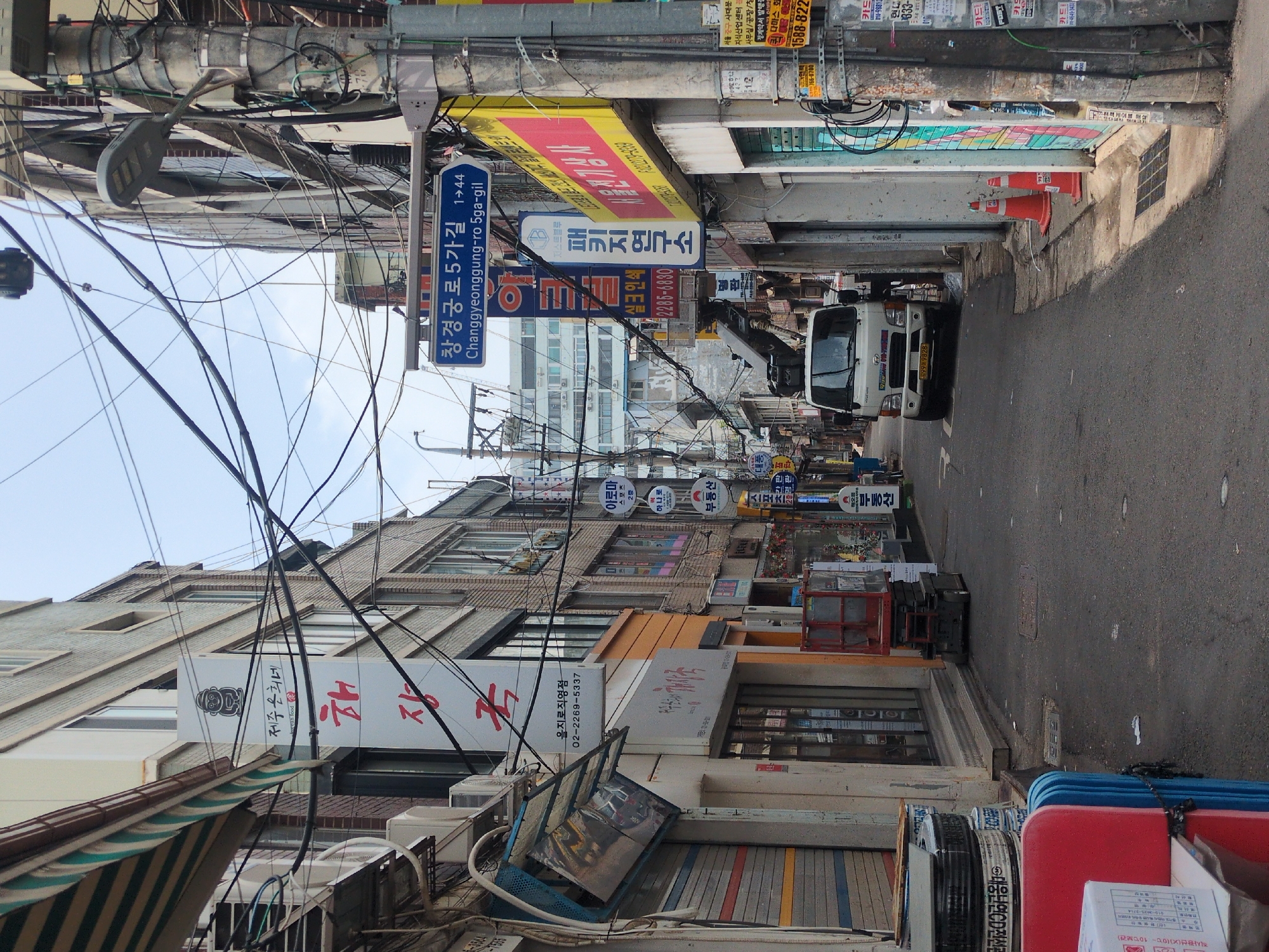 The image captures a narrow alleyway in Seoul, South Korea, bustling with activity and densely packed infrastructure. The street is lined with multi-story buildings, their facades cluttered with signs and various fixtures. A truck, with what appears to be a crane, is visible further down the alley. Overhead, a complex network of power lines and cables crisscrosses the space. The signs are mostly in Korean, advertising local businesses and services. Traffic cones are visible on the right side of the alley.