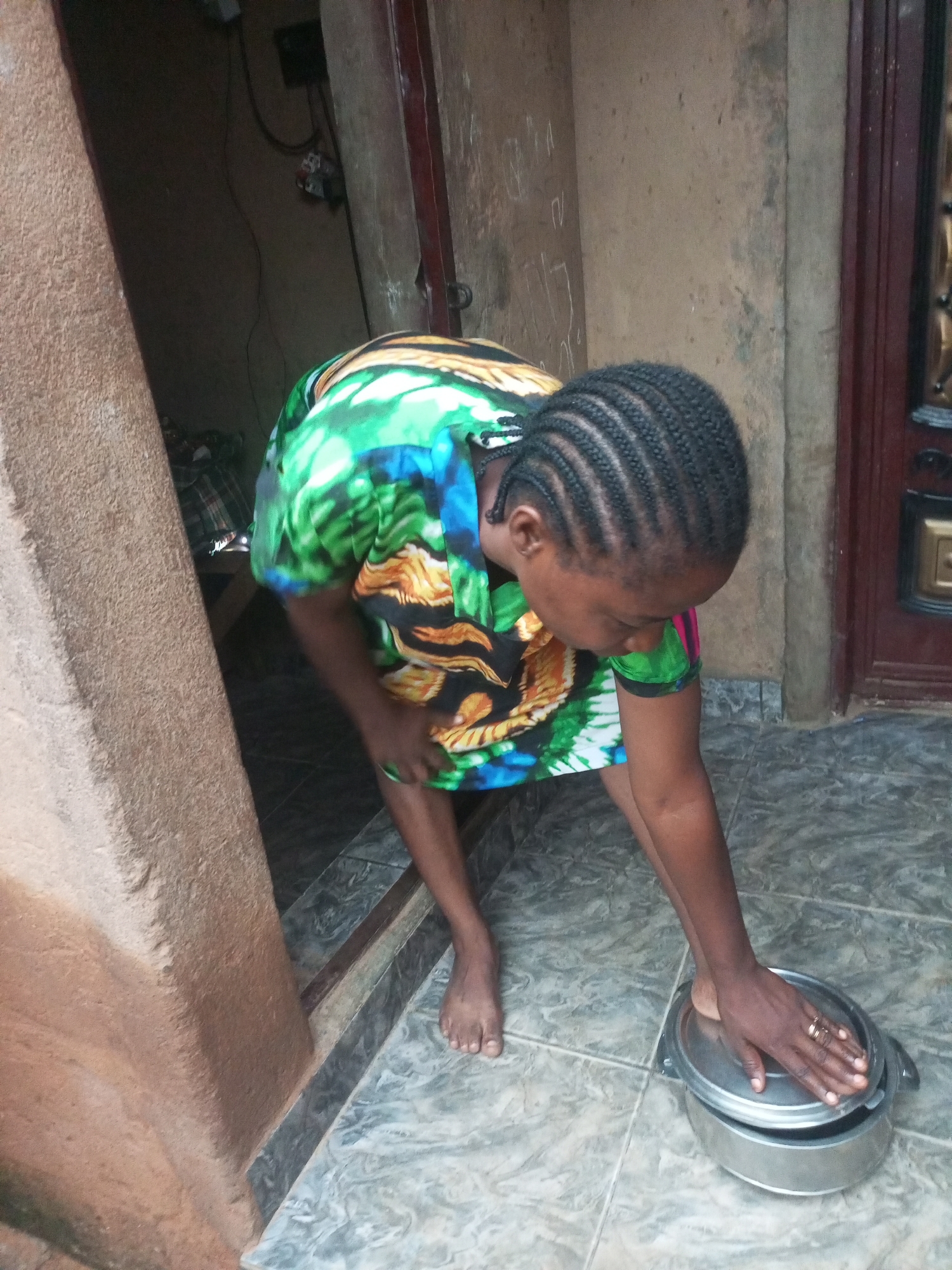A Nigerian woman with braided hair wears a brightly colored shirt and is bending over a silver pot on the floor. Her left hand is on the lid of the pot, and her right foot is also pressing down on it. She is standing on a tiled floor that appears to be outdoors, possibly in a courtyard or an alleyway. Behind her, a doorway leads into a dimly lit interior. The scene is located in Tempure, Nigeria.