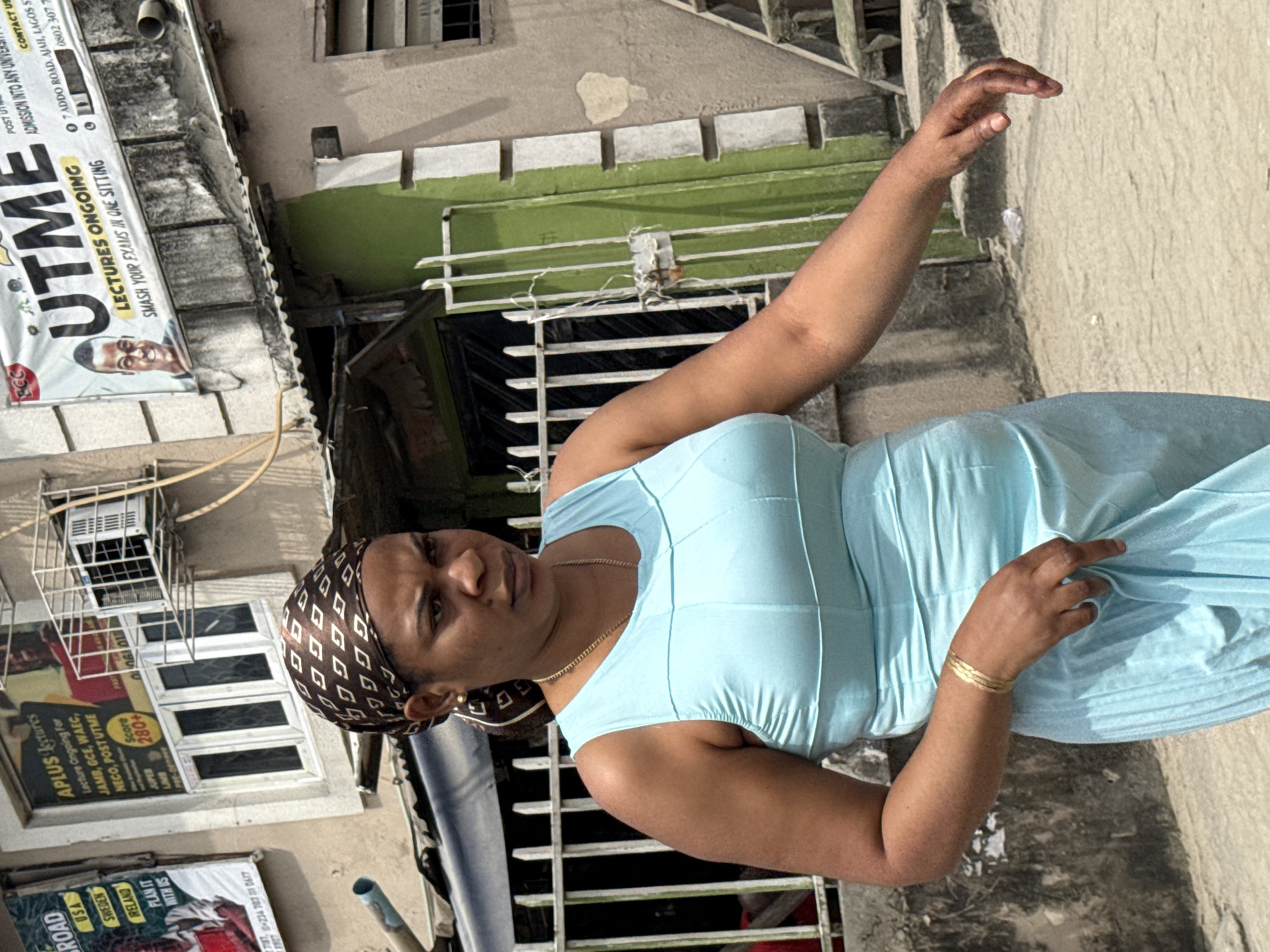 A woman wearing a light blue sleeveless dress and a brown patterned headscarf walks forward on a dusty street in Ado, Nigeria. She has a gold necklace and a gold bracelet on her left wrist. Behind her, a light green building has a metal gate and barred windows. A sign above the building reads "UTME LECTURES ONGOING SMASH YOUR EXAMS IN ONE SITTING." Another sign to the left advertises "APLUS Lectures" for JAMB, GCE, WAEC, NECO, and POSTUTME. The street is paved with concrete and sand, and other buildings and a staircase are visible in the background.
