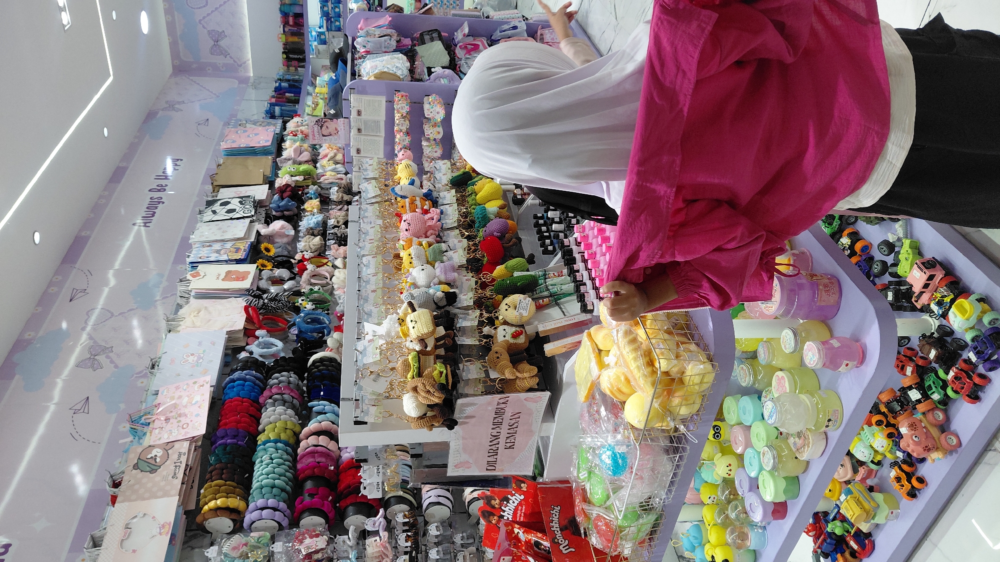 This image depicts the interior of a retail store, likely a gift shop or accessory store, situated in the city of Purwokerto, Indonesia. The store features brightly colored shelves displaying a variety of merchandise. On the upper shelves, numerous hair accessories, such as headbands and scrunchies in a spectrum of colors, are neatly arranged. Hanging displays showcase an assortment of plush keychains, many of which are cartoon or animal-themed, alongside smaller decorative items. Below these, shelves are laden with various small containers, possibly cosmetics or skincare products, and an assortment of small toy cars. In the foreground, a person wearing a white hijab and a bright pink outer garment is partially visible, facing away from the camera and appearing to browse or interact with the merchandise. The store's walls are decorated with pastel-colored murals featuring clouds, bows, and paper airplanes, with the text "Always Be Happy" prominently displayed. A sign with Indonesian text, "DILARANG MEMBUKA KEMASAN," meaning "DO NOT OPEN PACKAGING," is affixed to one of the displays. The lighting is bright, suggesting an indoor retail environment.