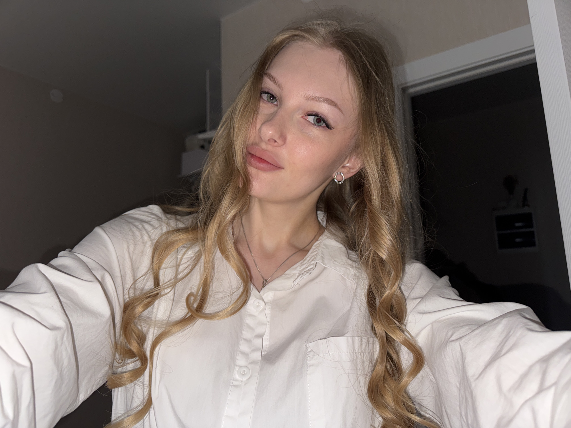 The media file is a close-up, selfie-style photograph of a young woman with long, wavy blonde hair. She is wearing a white collared shirt and a delicate silver necklace. She has light-colored eyes, and her makeup includes eyeliner and mascara, and her lips are painted a soft pink. She is looking directly at the camera with a slight smile, her expression appears calm and pleasant.
The background is dimly lit and appears to be an indoor setting. To the left, a dark wall is visible, and to the right, a doorway with a white frame is partially seen, suggesting a room or hallway. The lighting is low, creating a slightly dramatic effect with shadows, and the time of day is likely evening or night due to the low light conditions.
There are no visible objects or text that provide significant contextual information about the activity or location beyond it being an indoor setting. The overall impression is that of a casual self-portrait, possibly taken at home. The context provided indicates the location is Perm, Russia.