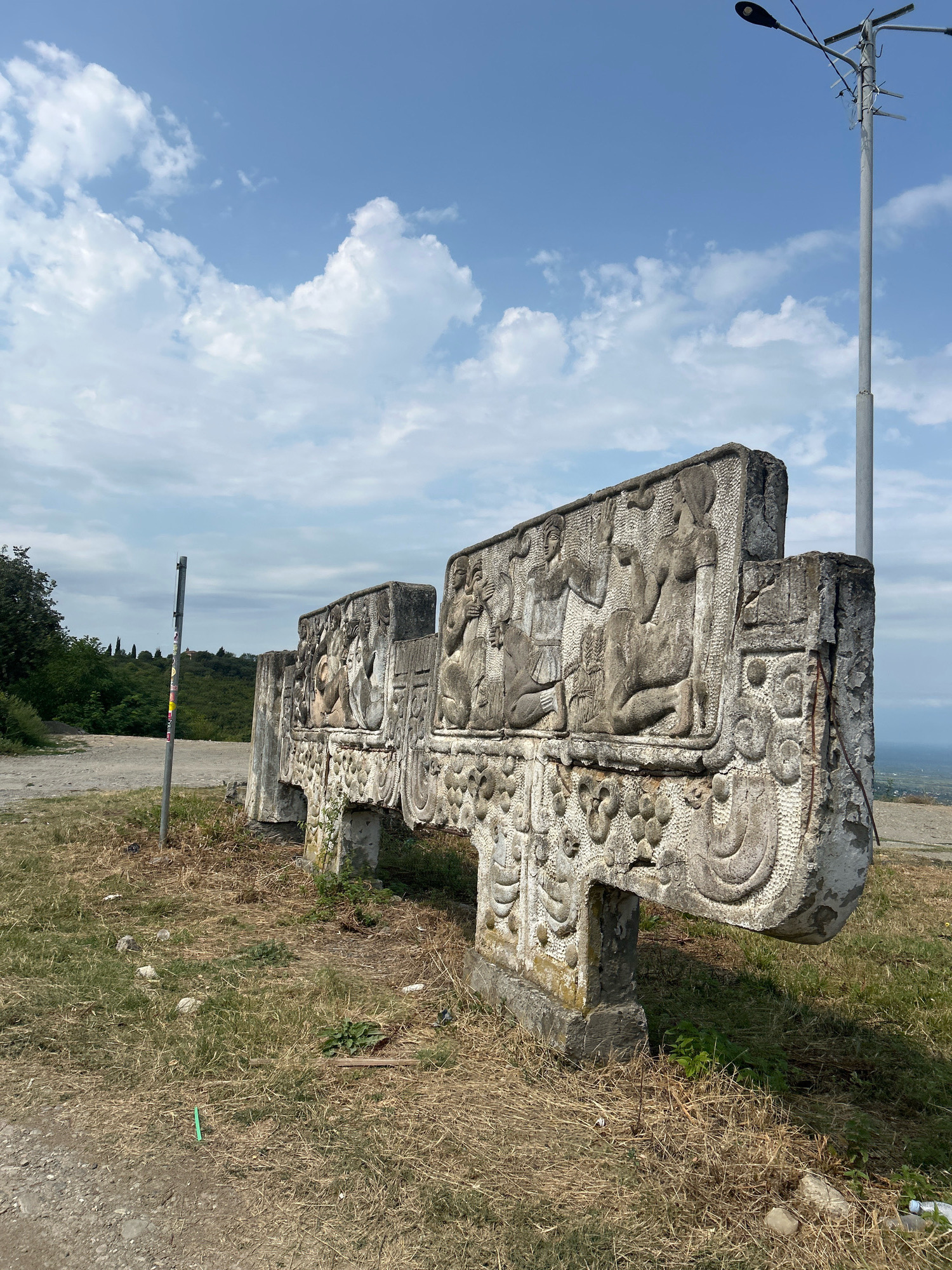 The image shows a stone monument in the city of Sighnaghi, Georgia, with a weathered appearance. The monument is characterized by intricate carvings that depict human figures and decorative patterns. It is situated outdoors on a grassy hill overlooking a landscape that appears to be a valley or plains, partially obscured by distant trees and hills. The sky above is blue with scattered white clouds. A modern street lamp stands tall to the right of the monument, and a slender metal pole with stickers is visible to the left. The ground around the monument is covered with dry grass, dirt, and some scattered debris. The monument itself appears to be made of concrete or stone and shows signs of age and exposure to the elements.