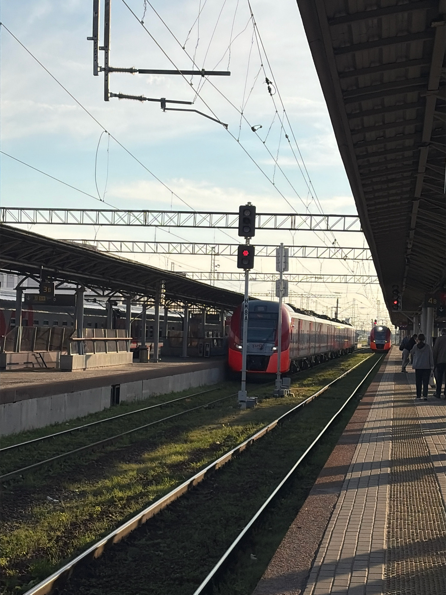 This image depicts a train station platform in Nizhny Novgorod, Russia. Two red and silver electric trains are visible, one closer to the foreground and another further down the tracks. The foreground train is stopped at a platform under a covered structure. A traffic signal with red lights is positioned near the train. Several people are standing on the platform in the background, some appearing to be waiting or walking. The platform is paved with tiles, and the tracks are bordered by grass. Overhead, power lines and supporting structures are visible against a pale blue sky with scattered clouds. The station appears to be active, with trains and people present.