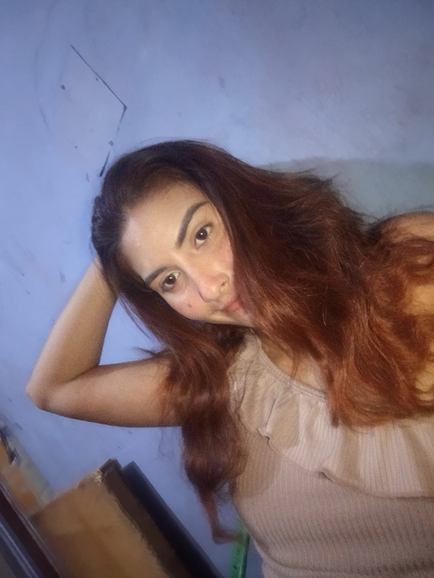 A person is indoors with a wall behind them. The person is wearing a tan, ribbed tank top. They have brown hair and brown eyes. One arm is raised above their head. There are scuff marks on the wall. There is a piece of furniture in the lower left corner of the image. The location is unknown and cannot be determined from the image.