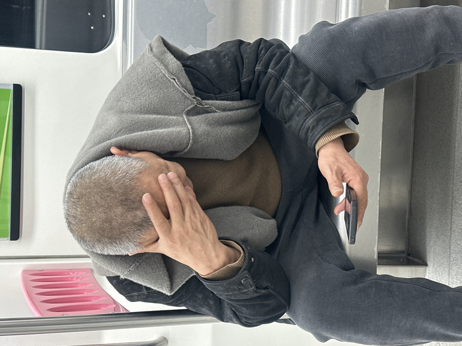 An adult male is seated on a metallic bench inside a subway train car in Xiema Subdistrict, China. He has short, graying hair and is dressed in a dark jacket, a grey scarf or cowl-neck garment, a brown long-sleeved shirt, and dark grey ribbed pants. The man's head is bowed, and his right hand covers his face. His left hand holds a black smartphone. The subway interior features white walls, a vertical white handrail, a curved pink partition, and a green display panel overhead.