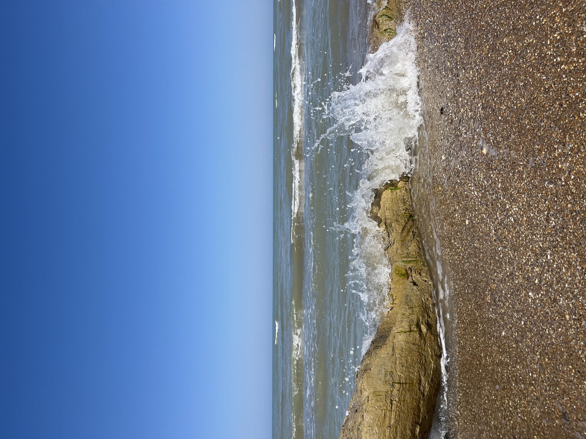 The image depicts a view of the Caspian Sea shoreline in Kaspiysk, Russia, under a clear, bright blue sky. The scene captures the point where the water meets the land, showcasing a small wave crashing against a rocky outcropping and a sandy beach. The beach is composed of coarse sand or small pebbles. There are no people visible in the image.