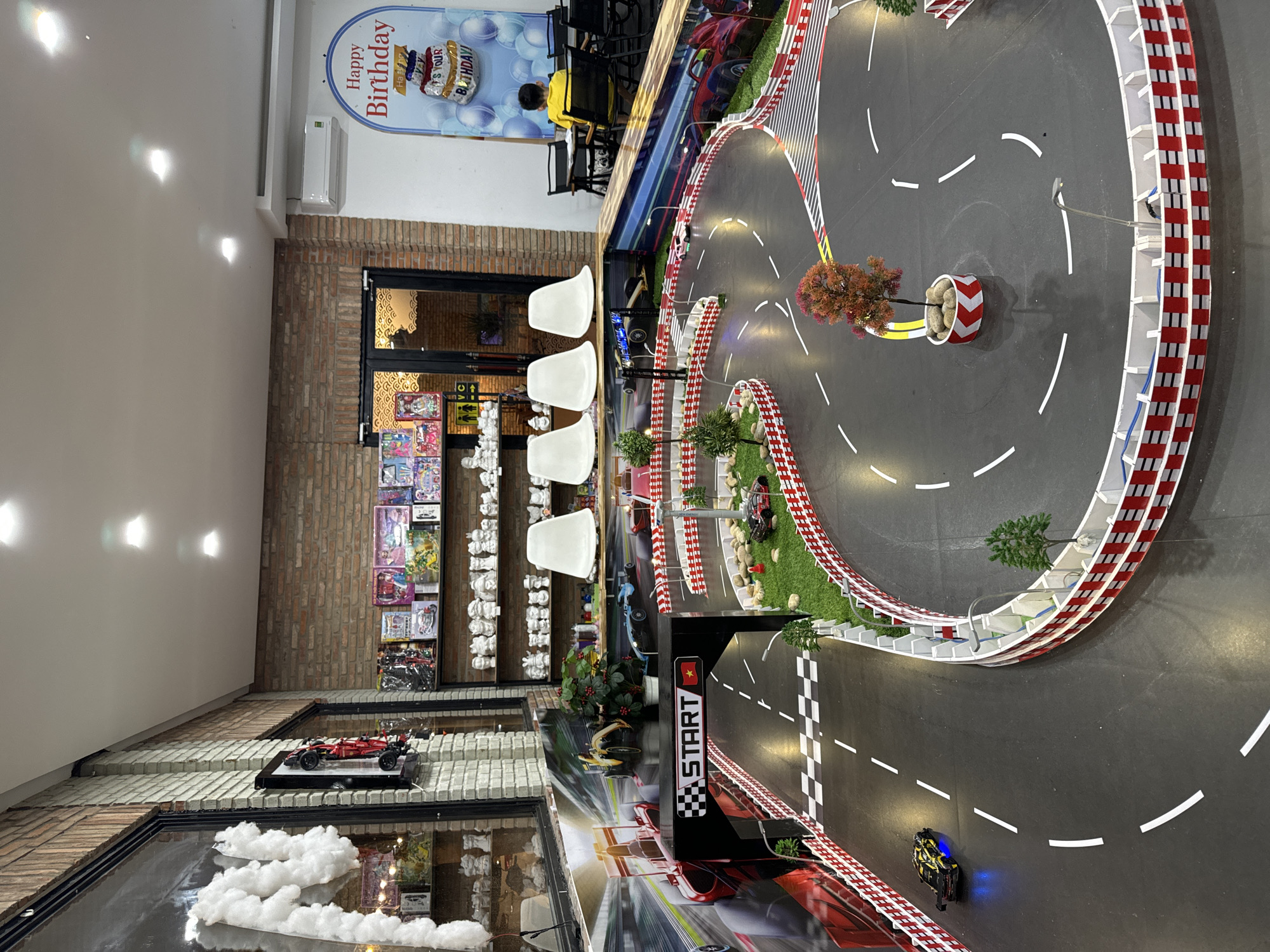 This media file captures an indoor scene in Phu Thuy Ward, Vietnam, depicting what appears to be an entertainment venue or cafe, dominated by a large, elaborate miniature race track for remote-controlled cars. The track features a dark gray asphalt surface with white lane markings, red and white checkered safety barriers, and interspersed green grassy sections adorned with small decorative trees and rocks. A prominent "START" gate displays a checkered flag pattern and the flag of Vietnam. Several miniature remote-controlled cars are visible on the track, including one with glowing blue lights in the foreground and a white car further along the course.
The setting includes brick walls and a white ceiling with recessed lighting fixtures. In the background, a "Happy Birthday" banner, featuring a cake graphic and the text "IT'S YOUR BIRTHDAY!", hangs on a wall, indicating the venue likely hosts celebrations. Below the banner, a child in a yellow shirt is seated at a table with black chairs, facing away from the camera. Another person, wearing a yellow top, is partially visible behind a track barrier. Closer to the foreground, a set of white, modern chairs are arranged around tables.
Along one brick wall, shelves are filled with numerous small, white ceramic figurines, possibly intended for painting, alongside various colorful packages that resemble books or craft supplies. In the lower foreground, a model red Formula 1 race car is displayed on a tiered brick structure. Additionally, fluffy white cloud-like decorations are visible near the bottom left corner. An air conditioning unit is mounted on a white wall section. The scene suggests a vibrant and engaging atmosphere for recreational activities.