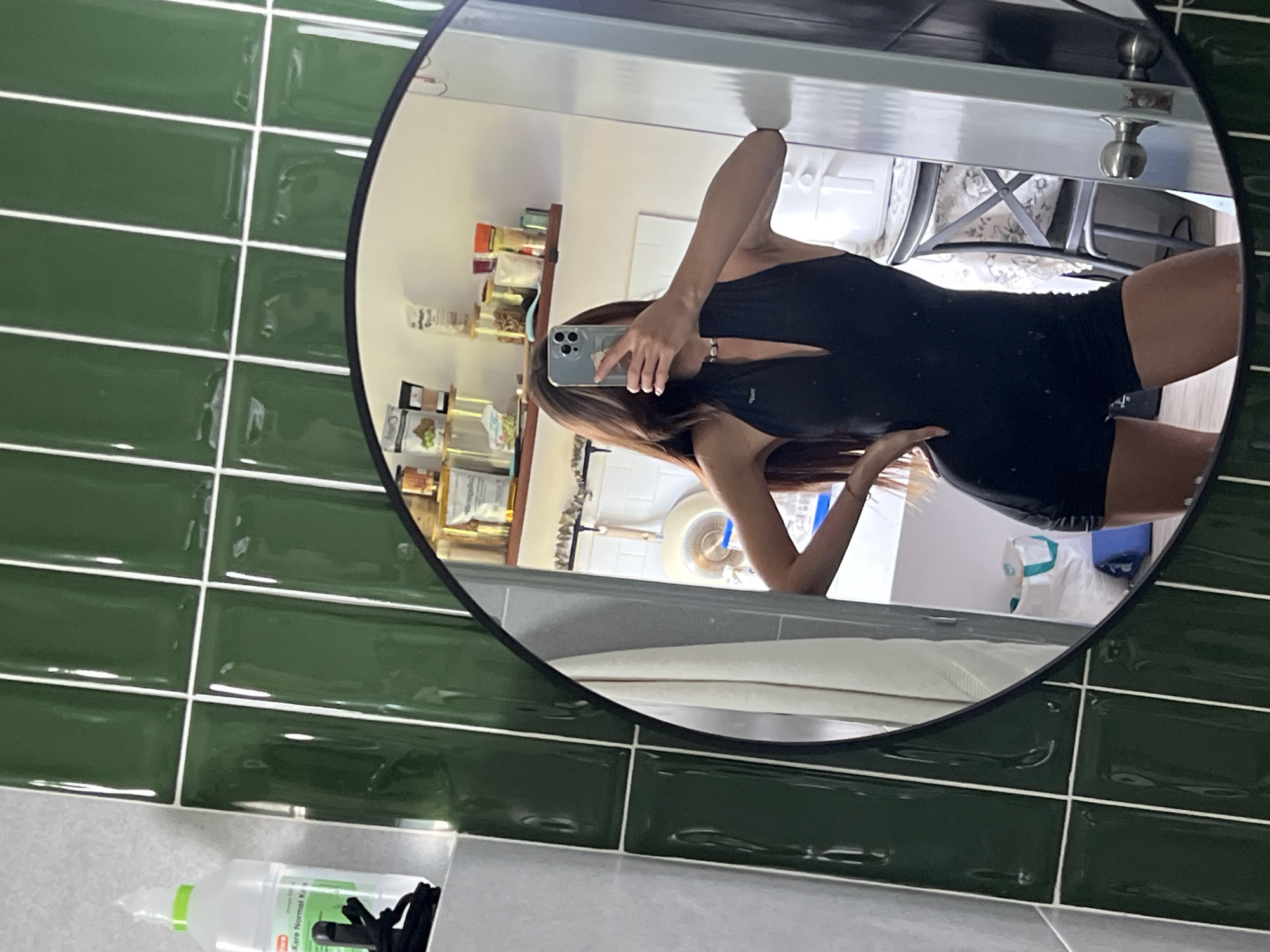 The image shows a mirror reflecting a person taking a selfie with their phone. The person is wearing a black outfit. The reflection shows a kitchen with shelves of jars and containers, as well as a chair and other kitchen items. The mirror is mounted on a wall with dark green tiles. A bottle of Kare Normal Kiss spray is in the corner of the image. The location is Unknown, Unknown.