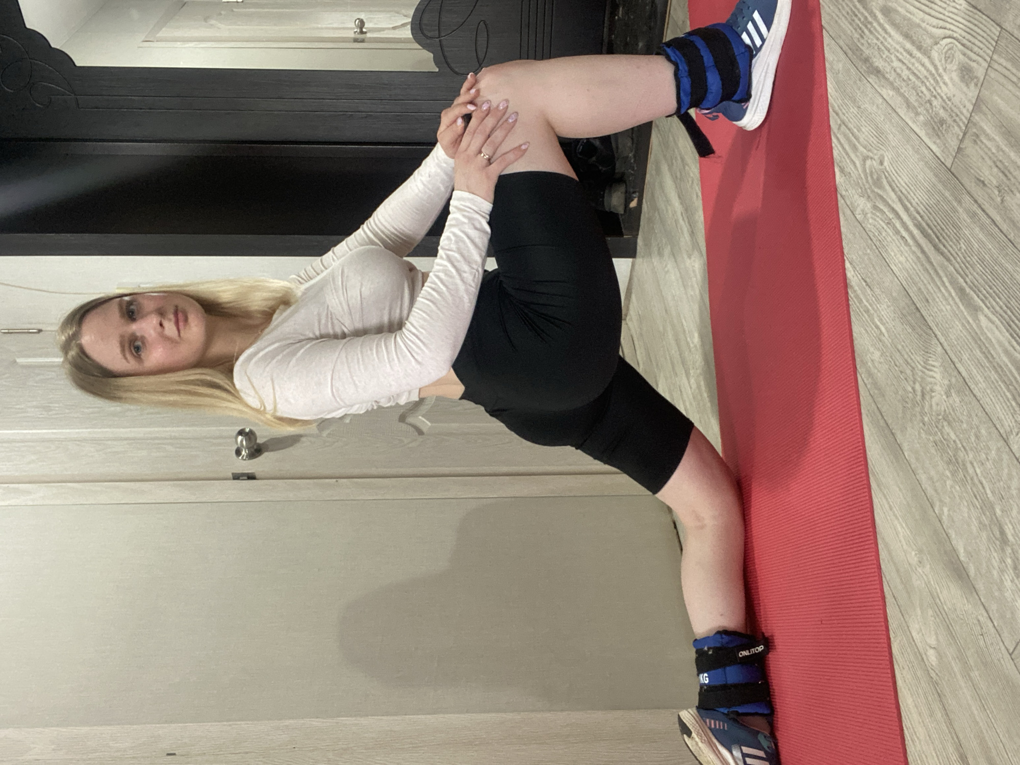 A young adult woman with long blonde hair is performing a stretching exercise indoors, on a red yoga mat. She wears a white long-sleeved top and black athletic shorts. Her legs are positioned in a split, with ankle weights strapped to her sneakers. The setting appears to be a room with a light-colored door and dark wood furniture. The floor is covered in a light gray wood-patterned material. I cannot verify the location in the city of Chaikovsky, Russia.
