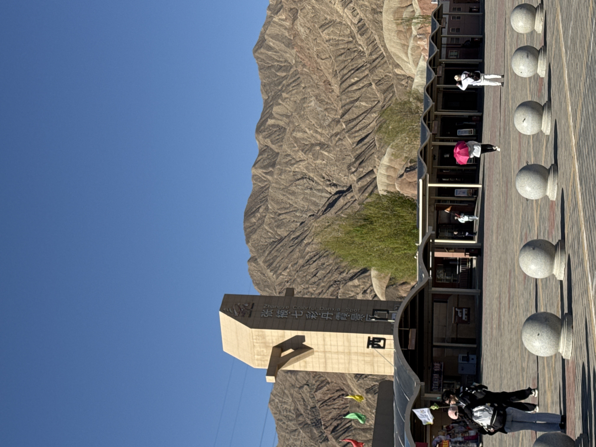 This image captures a vibrant scene at the Zhangye Colorful Danxia Scenic Area in Nantai, China, on a clear, sunny day. The background is dominated by majestic, rugged mountains showcasing the distinctive layered geological formations of Danxia landforms. Above, a bright, expansive blue sky suggests fair weather.
In the foreground, a wide, paved plaza or walkway stretches across the scene, lined on the right by a row of decorative spherical stone bollards. Along the left side of the plaza, modern buildings with curved roofs provide sheltered spaces, possibly housing visitor amenities. Several people are present on the plaza, engaging in leisurely activities typical of tourists. One individual in white stands near the buildings further back, while another walks towards the right holding a striking bright pink umbrella. Closer to the viewer, two more people are seen walking together, one with a backpack.
A significant stone monument or entrance sign is prominently featured on the left, displaying both Chinese characters and English text. The visible English text clearly identifies the location as "Zhangye Colorful Danxia Spot". Below this, colorful flags in yellow, green, and red add a touch of vibrancy to the scene. The overall impression is that of a popular and well-maintained tourist destination, bustling with visitors enjoying the unique natural beauty.