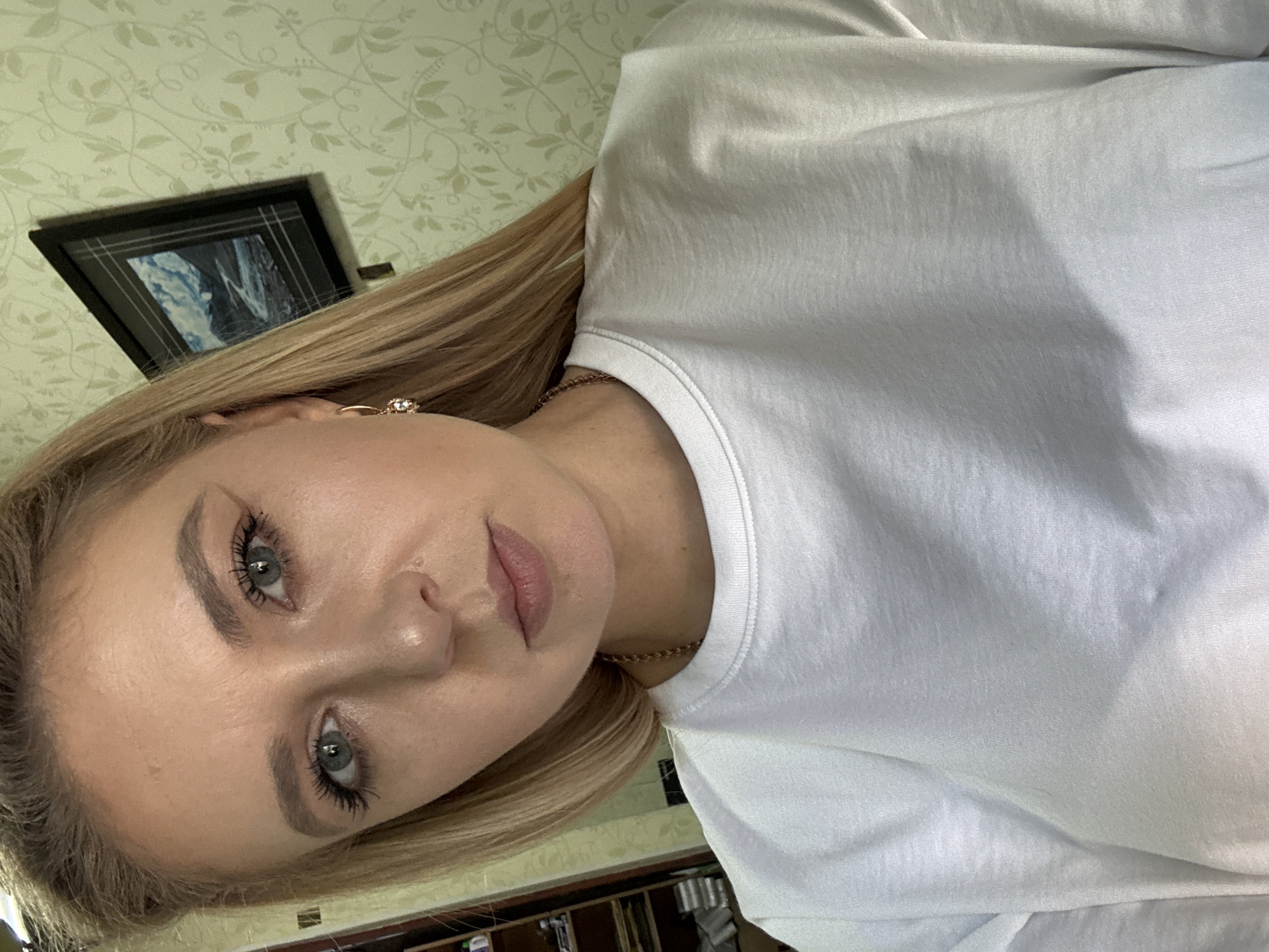 A medium shot captures a blonde woman from the chest up, looking directly at the camera. She has light blue eyes, light makeup, and her hair is parted on the left and falls over her right shoulder. She is wearing a white t-shirt and a gold chain necklace. Her left ear, visible, has a gold earring with a small gem. In the background, to the right, a framed picture of a landscape hangs on a patterned wallpaper. To the left of the woman, a bookshelf is partially visible, filled with books and other items. The overall setting appears to be indoors, in what seems to be a domestic environment.