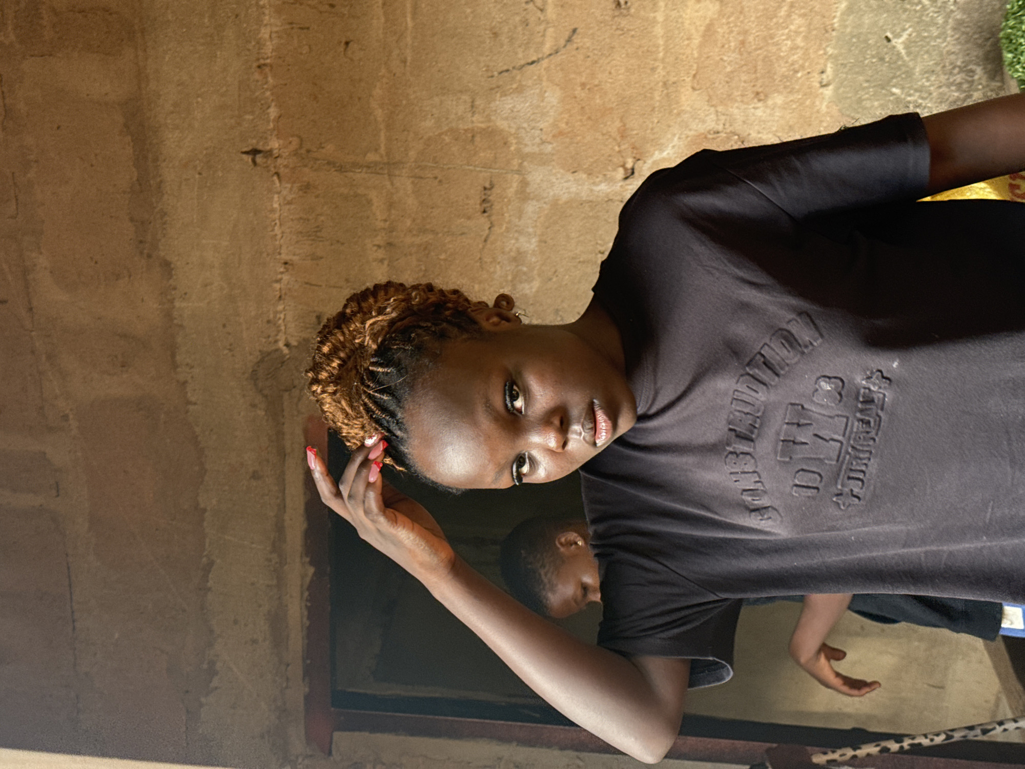 The image presents a portrait of a young Black woman, centrally positioned and looking directly at the viewer. She has intricately braided hair, featuring warm brown tones, and her left hand is raised near her face, revealing bright red or pink painted fingernails. She is wearing a dark, possibly black, short-sleeved t-shirt with embossed or raised lettering across the chest. The word "EDITION" is clearly visible, along with other partial, indistinct letters and symbols. Her expression is neutral to slightly serious.
In the blurred background, another person, likely younger, is partially visible to the main subject's right, also wearing a dark t-shirt. Their head and upper body are discernible but out of focus.
The setting appears to be indoors, against a textured, light brown or beige wall that looks like concrete or a similar rough material. Faint dark markings and lines are visible on the wall surface. The lighting is soft and natural, suggesting it is daytime. The overall scene captures a casual moment in Eziudo, Nigeria.