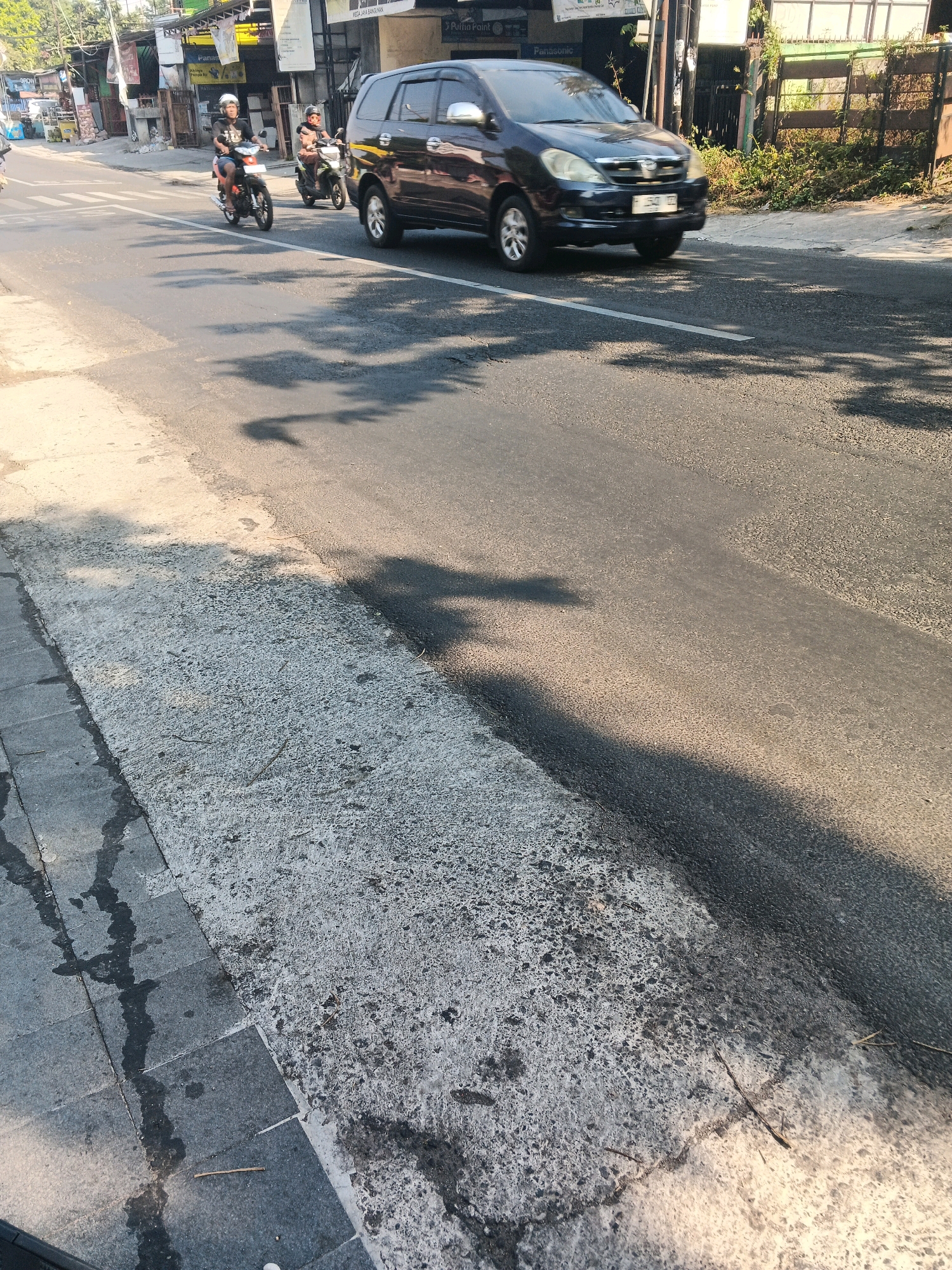 The image shows a street scene in Ungaran, Indonesia, during daytime. Two individuals are riding motorcycles on the asphalt road. The first motorcycle is black with a rider wearing a white helmet and a dark shirt. The second motorcycle is green and is being ridden by a person wearing sunglasses and a dark helmet. A dark-colored SUV is also visible on the road, appearing to be in motion. The street is lined with buildings and vegetation on the right side, and a pedestrian walkway with some water stains is on the left. Shadows of trees are cast across the road, indicating a sunny environment. The road surface shows some wear and tear.