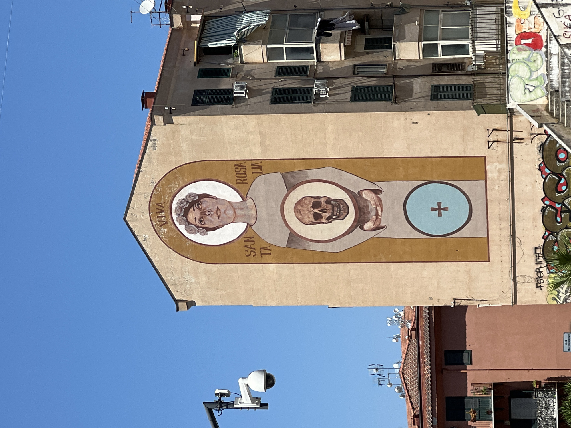 This media file captures a mural painted on the side of a building, likely in Palermo, Italy, given the location context. The mural depicts a stylized religious icon within a long, rectangular frame. At the left of the frame is a haloed head with curly hair and a serene expression, identified by text as "SANTA ROSALIA." To the right of the head, the mural features a skull, a common symbol of mortality and penance, held by hands that emerge from flowing robes. Further to the right, within a blue circle, is a red cross. The overall scene is bathed in bright sunlight, suggesting a clear day and likely daytime.
The building itself appears to be multi-story, with visible windows and shutters. A satellite dish and antennas are mounted on the roof, common in urban environments. The wall next to the mural is covered in graffiti. Below the mural, a security camera is mounted on a pole, overlooking the street. The bottom of the image reveals more of the urban landscape, with tiled roofs and other buildings, and a street sign that reads "ZONA TRAFFICO LIMITATO" (Limited Traffic Zone). The mural's imagery, featuring Santa Rosalia, the patron saint of Palermo, and symbols of faith and mortality, suggests a cultural or religious context specific to the city.