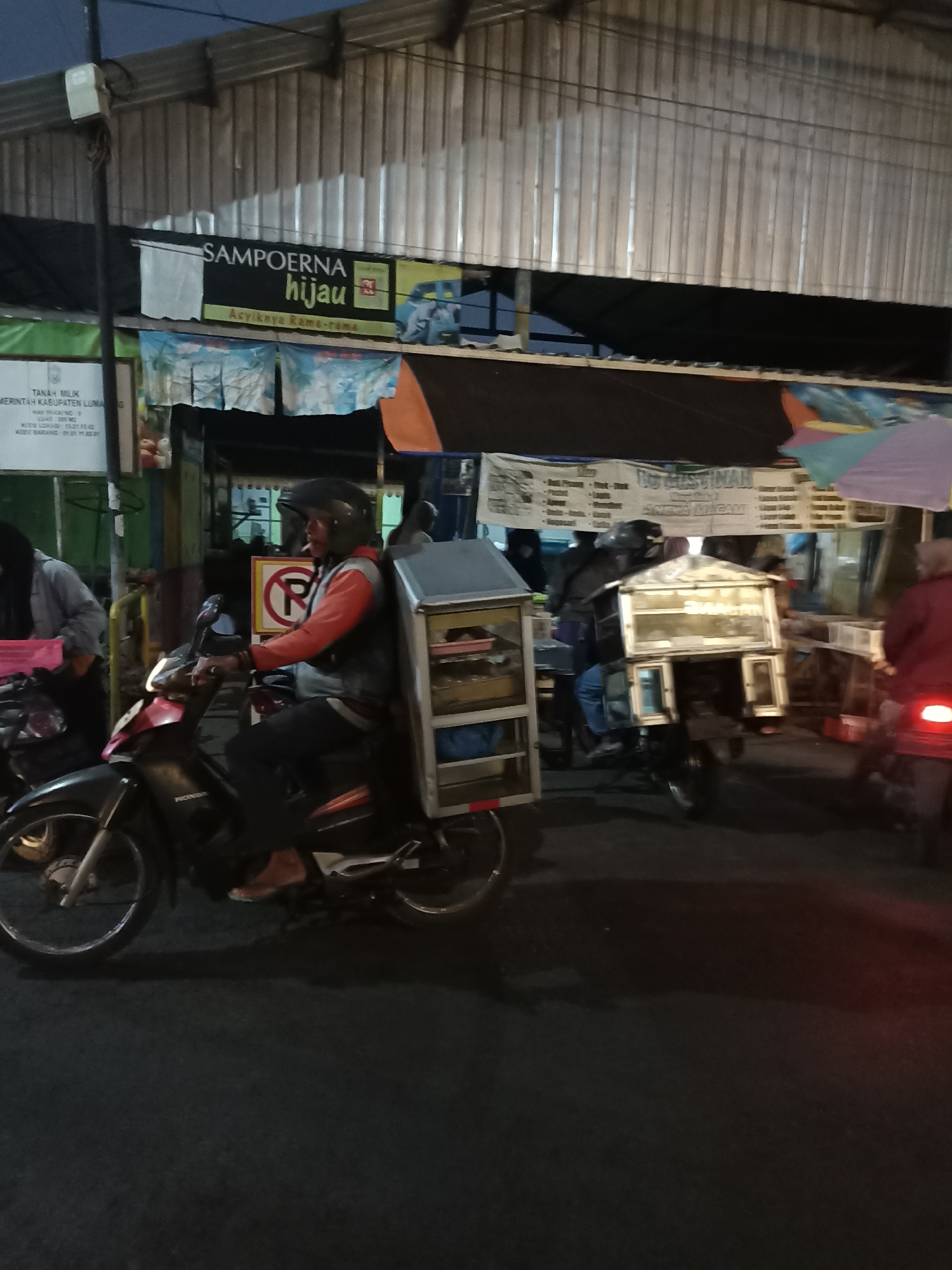 The image shows a nighttime scene on a street in Ditotrunan, Indonesia. In the foreground, a person is riding a motorcycle. The motorcycle is red and black and has a metal cargo box attached to the back, which is illuminated. The rider is wearing a black helmet and a red and gray jacket, and they are holding a cigarette. Behind the rider, there is a sign that says "SAMPOCRNA HIJAU Asyiknya Rama-Rama". Further back, there is another motorcycle with a similar cargo box. There are also several stalls or shops with banners and signs. The street is paved and appears to be dark, with artificial lighting from the shops and signs. The overall impression is of a busy street market or food stall area at night.