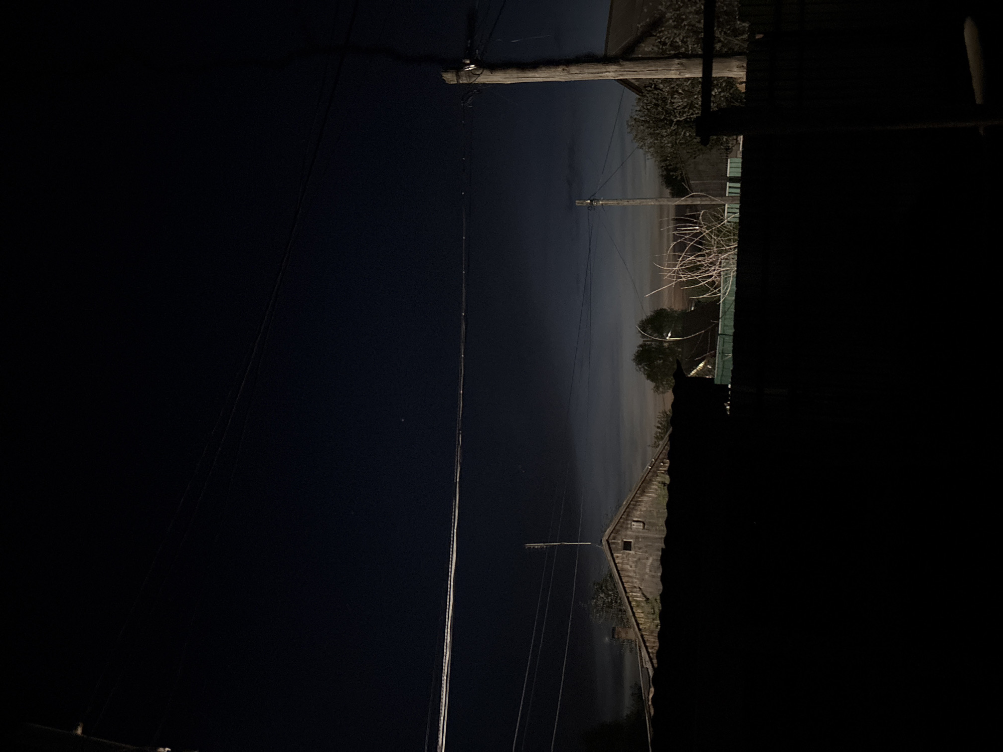 A dark, underexposed, and vertically oriented image taken at night in Фоки, Russia, shows a view between buildings or fences, with telephone poles and wires visible against a dark sky. Some foliage is present in the midground, lit dimly. The foreground is primarily obscured by deep shadow. The image's location within the city is inferred due to the presence of utility infrastructure (telephone poles and wires).
