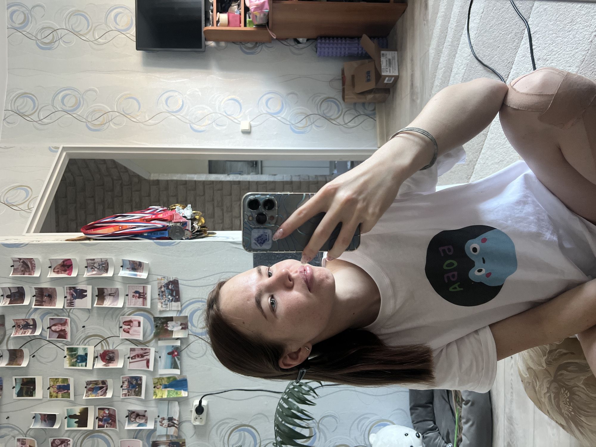 The image is a selfie taken in a room in Chaikovsky, Russia. A young woman with long brown hair is holding a smartphone up and facing the camera. She is wearing a white t-shirt with a blue frog design. Her left knee appears to be wrapped in a bandage.
Behind her is a wall covered in photos, a doorway leading to another room, and a glimpse of the room's decor. The room features patterned wallpaper, a television mounted on the wall, and a shelf with various items. Other objects in view include a grey couch, a plant, and a stuffed animal. The lighting appears to be natural.