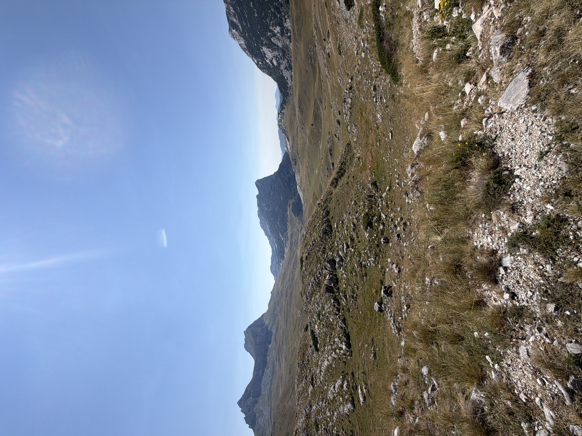 The image depicts a rugged, mountainous landscape under a clear blue sky. The terrain is characterized by rolling hills covered in dry grass and scattered rocks, with more imposing, rocky mountain peaks in the background. The light suggests it is daytime, likely mid-morning or early afternoon, with a bright sun creating a slight lens flare in the upper left quadrant. The vegetation appears sparse and adapted to a drier climate, with predominantly golden-brown grasses. There are no visible people, structures, or animals. The overall scene conveys a sense of natural, undeveloped wilderness. The location is identified as Dubrovsko, Montenegro.