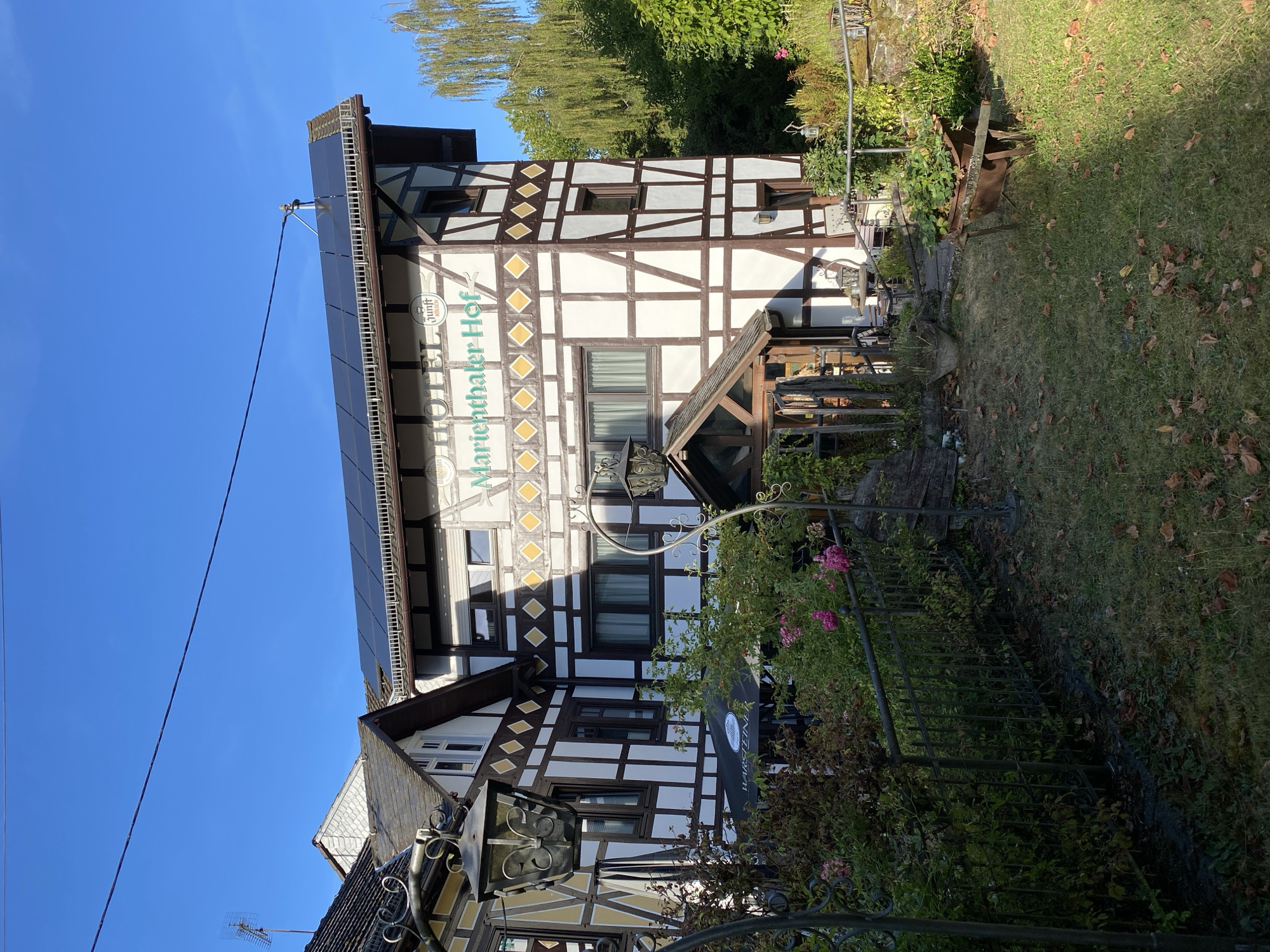 The image captures a view of the "Hotel Marienthaler Hof" located in Seelbach bei Hamm (Sieg), Germany. The hotel features traditional half-timbered architecture with white stucco and dark brown wooden beams, and a distinct diamond pattern on the upper sections of the walls. Solar panels are installed on the roof of the building. In the foreground, there are decorative lanterns, a black umbrella with text indicating "Warsteiner", and lush green bushes with pink flowers. A metal fence runs along the side of the property, separating the garden area. The ground is covered with grass and fallen autumn leaves. To the right, a rusty wheelbarrow filled with plants is visible in a grassy area. In the background, mature trees with green foliage contribute to the natural setting. The sky is clear and blue, suggesting a sunny day. There are no people visible in the image.