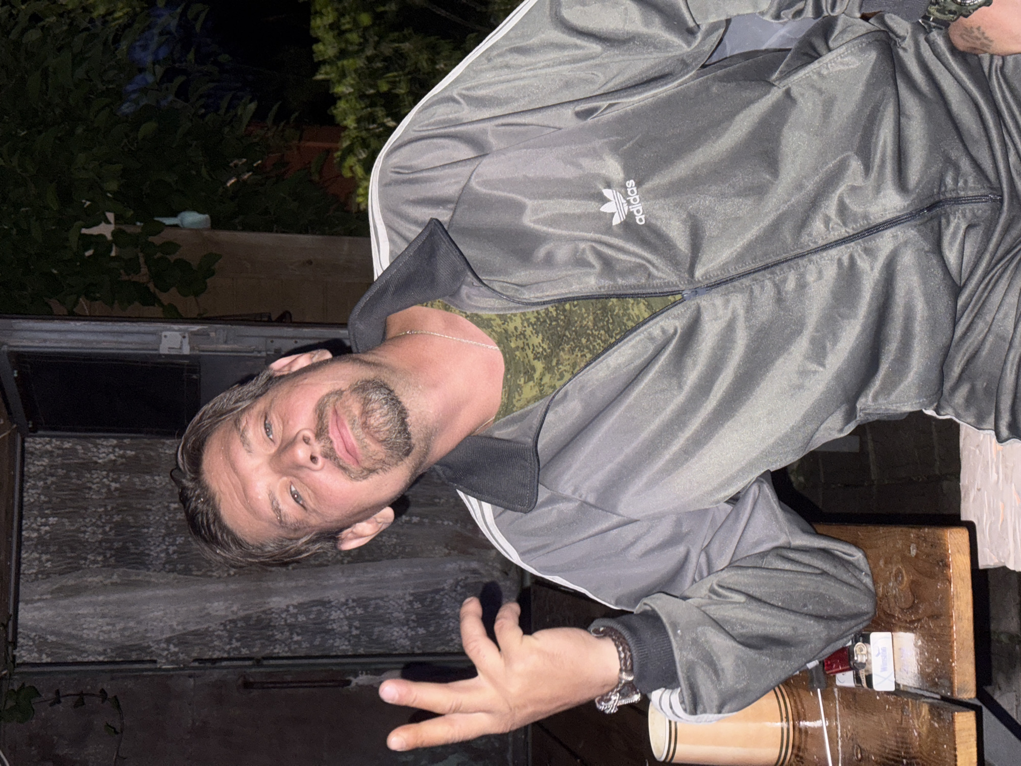 In the city of Автомобилист-1 Снт, Russia, a man is captured from the chest up, seated at a wooden table outdoors at night. He wears a grey Adidas tracksuit with white stripes on the sleeves and collar, and a camouflage t-shirt underneath. His right hand is raised, with fingers extended in a gesture. A silver chain with a pendant is visible around his neck. On the table are a beer mug, a pack of Winston cigarettes, and two lighters. The background shows a partially open door and lush green foliage.