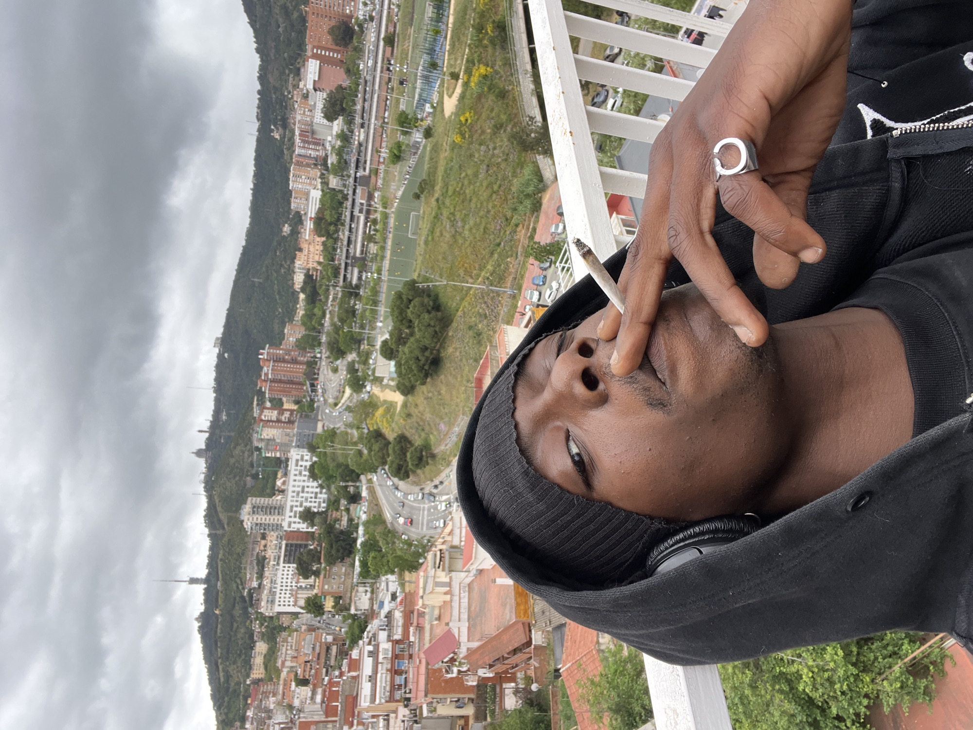 A man wearing a black hoodie and a beanie is smoking a cigarette on a balcony. He has headphones around his neck and a silver ring on his right hand. The balcony overlooks a cityscape with various buildings, trees, and a football field. The background shows a mountain and a cloudy sky. The location is Barcelona, Spain.