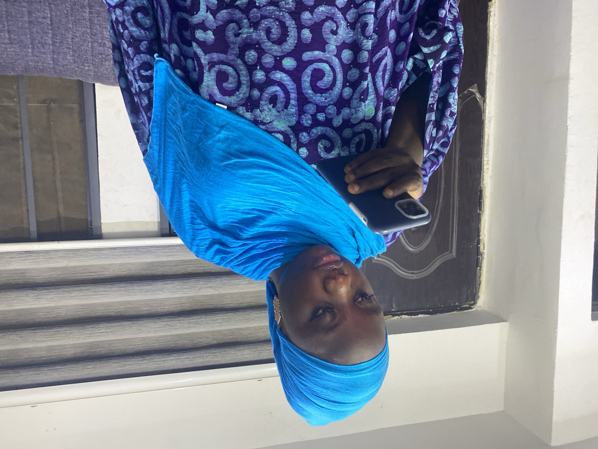 The image, taken in Abuja, Nigeria, features a woman captured from the chest up in an indoor setting. She has dark skin and is wearing a vibrant blue head covering that drapes over her left shoulder, and a long-sleeved dress with a distinctive purple and light blue/green swirl pattern. A single gold-toned earring is visible on her left ear. She holds a dark-colored smartphone, likely an iPhone due to the camera bump, in her right hand, with its screen facing away from the viewer.
The background reveals an indoor space. To the left, a window is fitted with horizontal blinds displaying alternating light and dark gray stripes. To the right, a section of a dark wooden door with a sculpted, arched panel is visible. The surrounding walls are a plain light color, possibly white or off-white.
The woman is looking directly at the camera with a neutral expression, suggesting she is posing for the photograph. The lighting is consistent with indoor artificial light or diffused daylight, making the exact time of day difficult to determine. No legible text is present anywhere in the image.