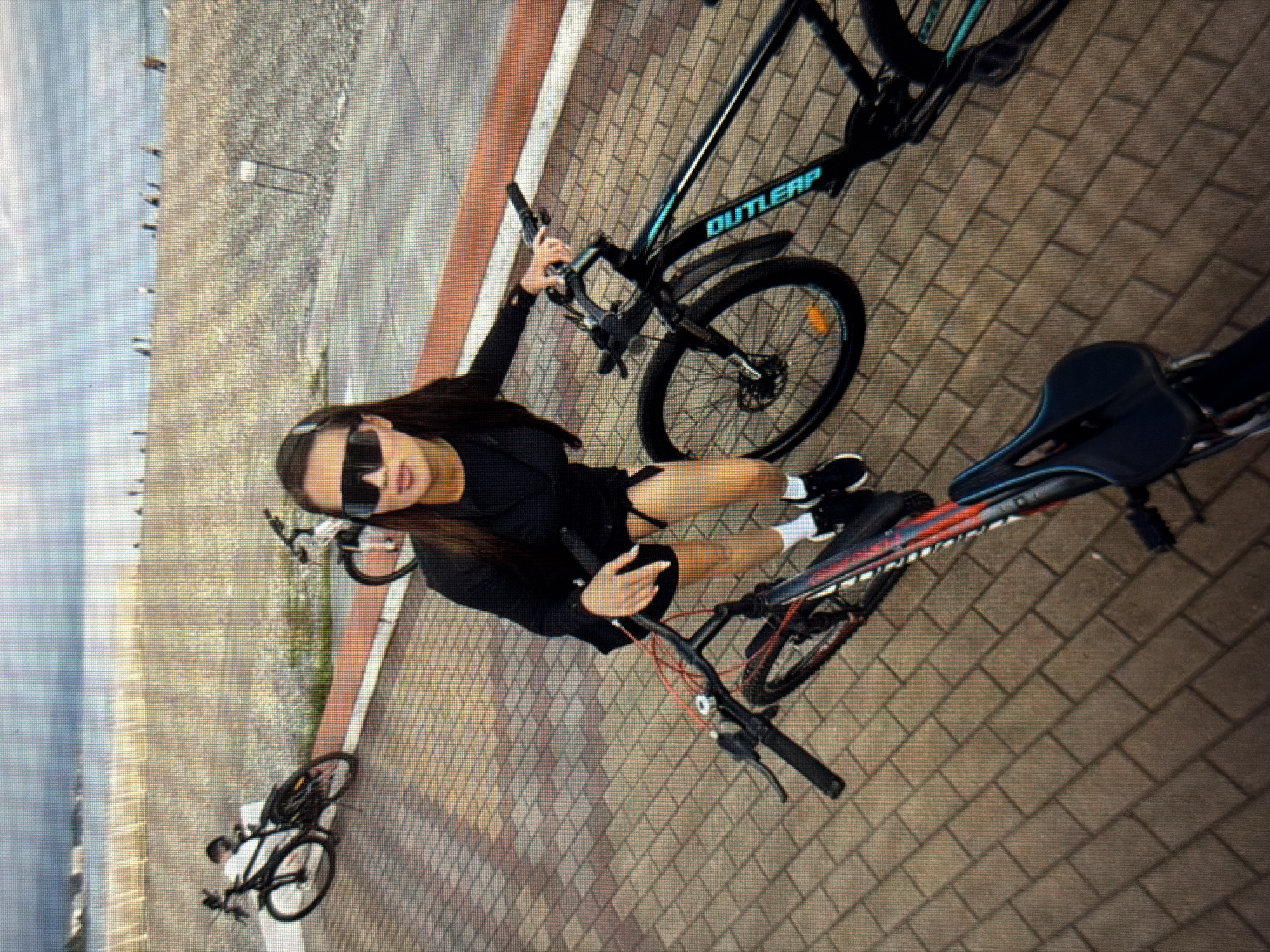 A young woman with dark hair and sunglasses is standing on a brick pathway near a beach. She is wearing a black jacket and skirt, white socks, and black sneakers. She is holding the handlebars of a black bicycle. In the foreground, another bicycle is visible. In the background, there is a beach with people and the ocean. The sky is overcast. The scene appears to be in the city of Sirius, Russia.