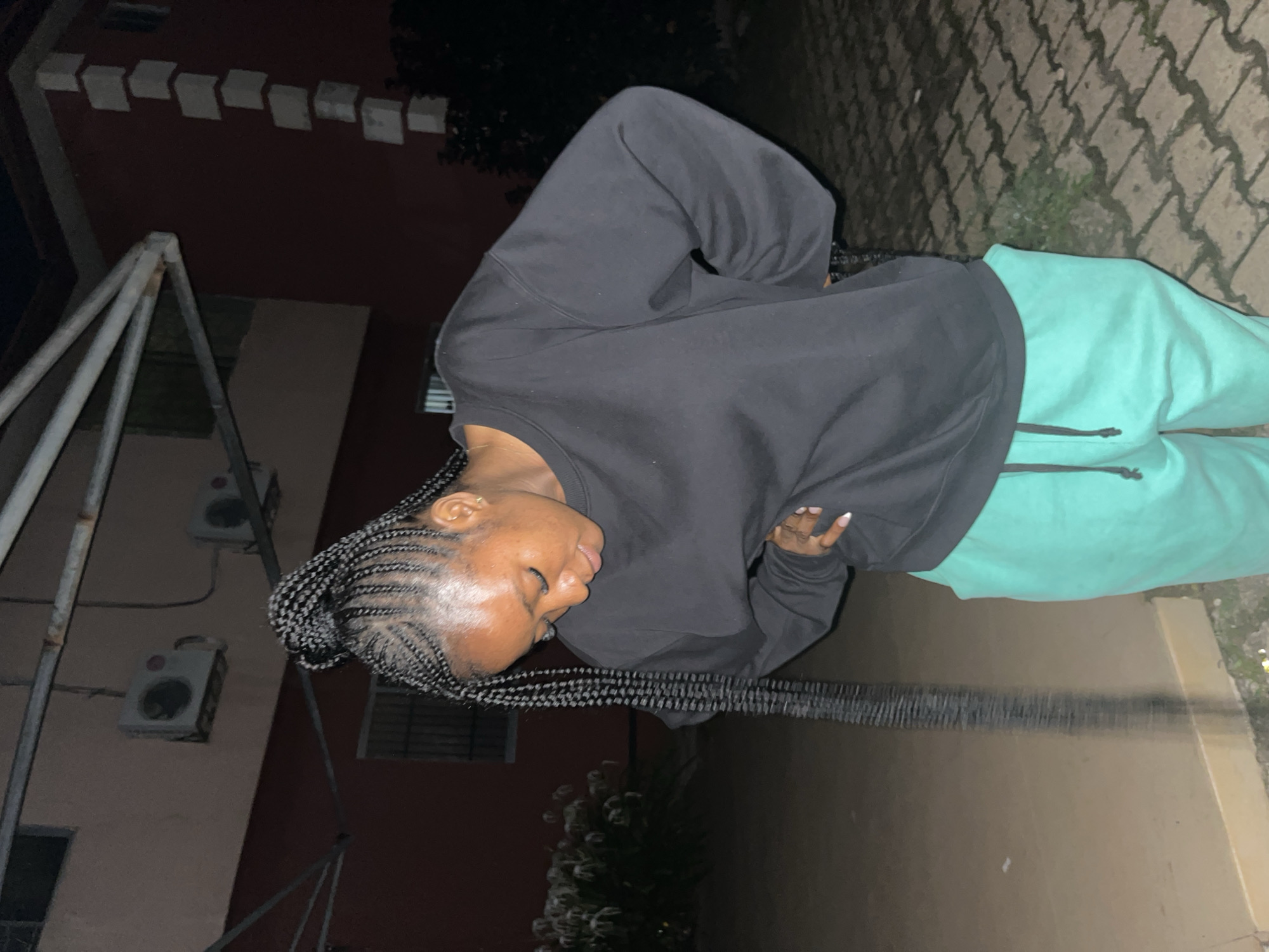 A young Black woman with braided hair is standing outdoors at night, looking down at her midsection. She is wearing a black sweatshirt and bright teal sweatpants. The background shows a building with red walls, air conditioning units mounted on the wall, and a paved ground. There are some plants visible in the foreground. The image appears to have been taken in Jos, Nigeria.