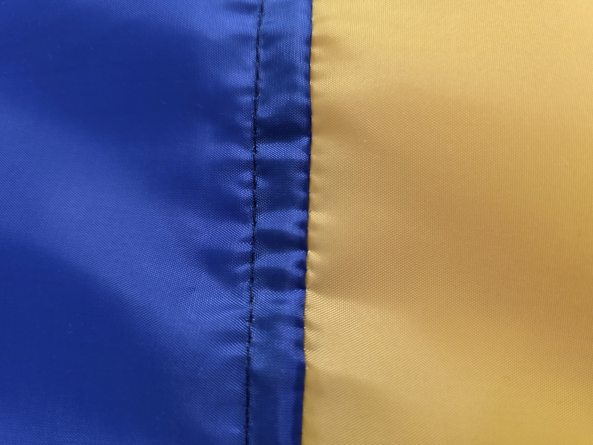 The image displays a close-up, abstract view of the Ukrainian flag. The top half is a vibrant blue, and the bottom half is a rich golden yellow. A thick, dark seam runs horizontally across the center, dividing the two colors. The fabric appears to be a synthetic material, with a slightly textured weave visible upon close inspection. The lighting suggests a natural light source, casting subtle shadows and highlights on the rippling fabric. There are no discernible landmarks, people, or specific locations visible that would indicate a precise spot within the city of Kyiv, Ukraine. The image is purely a representation of the flag's texture and colors.