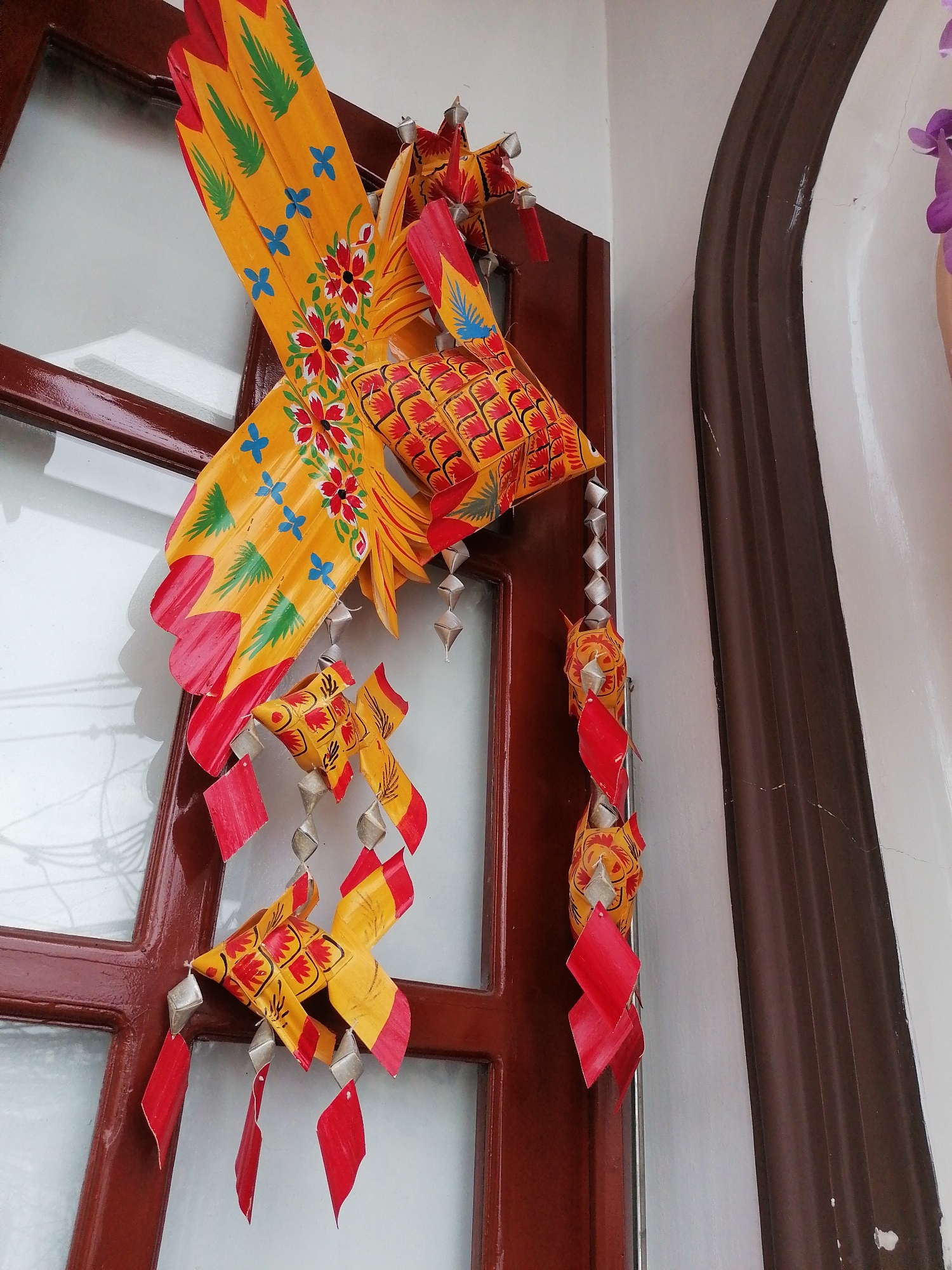 A vibrant, hand-painted paper decoration hangs from a dark brown wooden door frame in what appears to be a residence in Da Lat, Vietnam. The decoration is predominantly yellow and red, shaped like a stylized bird with fish-like elements. The bird has a large, outstretched wing adorned with hand-painted floral and foliate designs in red, green, and blue. Beneath the main bird figure, there are several smaller fish-shaped elements dangling, connected by a string of silver-colored, diamond-shaped metal pieces, adding a sense of movement. These smaller fish are also painted with similar yellow and red patterns and have red diamond-shaped appendages. The background includes a white wall with a dark brown, vertically-oriented architectural detail. The window behind the door frame reflects light, suggesting an outdoor environment. The overall image suggests a traditional Vietnamese craft, likely a souvenir or a decoration used for cultural celebrations.