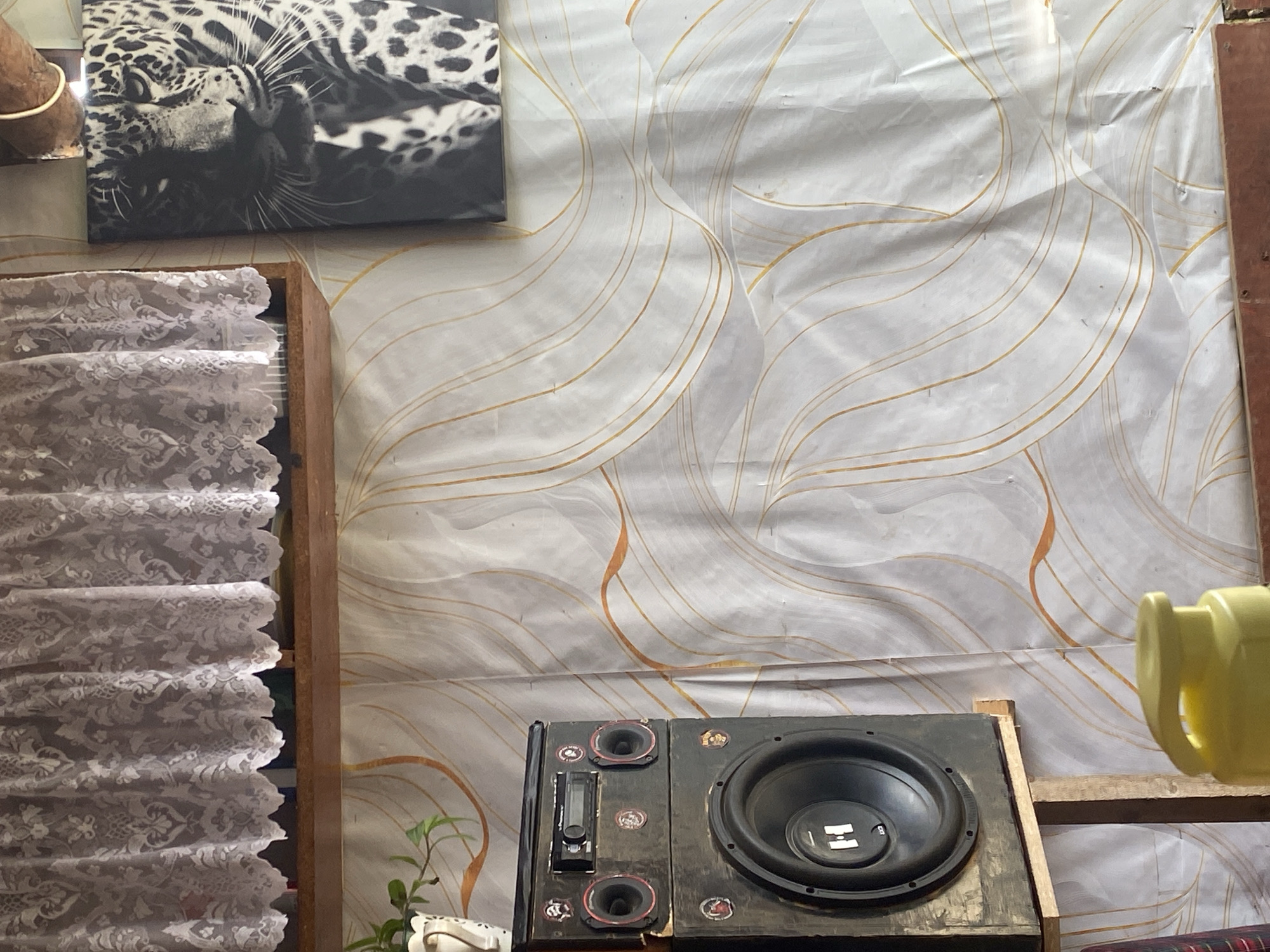 The image depicts an indoor scene, likely a room, with a patterned wallpaper featuring flowing lines in white and orange. A white lace curtain hangs across the top portion of the frame, covering a wooden shelf. To the right, a black and white framed photograph of a leopard's face is displayed. On the left side of the image, a stacked speaker system is visible, with the top unit containing a car stereo. The larger speaker below has a black cone with a white "H" logo. The speaker system is mounted on a simple wooden stand. In the foreground, a yellow plastic dispenser with a curved handle is partially visible. The overall impression is of a somewhat cluttered living space. The location is identified as Borodulino, Russia, although this context is not visually apparent from the image itself.