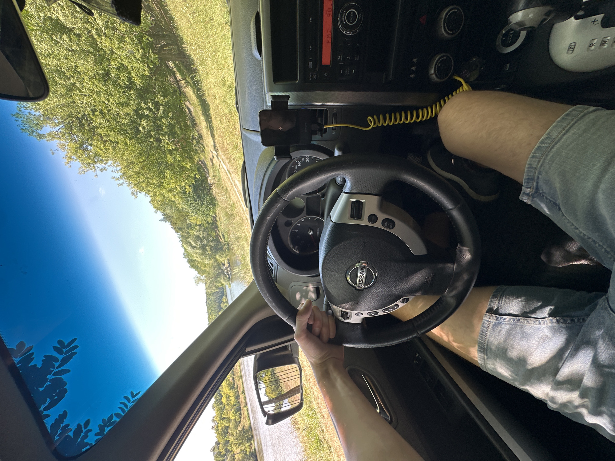 The image shows the interior of a Nissan X-Trail driving on a dirt road next to a river. The driver's hands are on the steering wheel, and their legs are visible. The dashboard has a phone mount with a charging cable. The exterior view shows a river and trees, indicating a natural, possibly rural, setting. There is no indication of the location being the city of Makiv, Ukraine, or any other specific location.