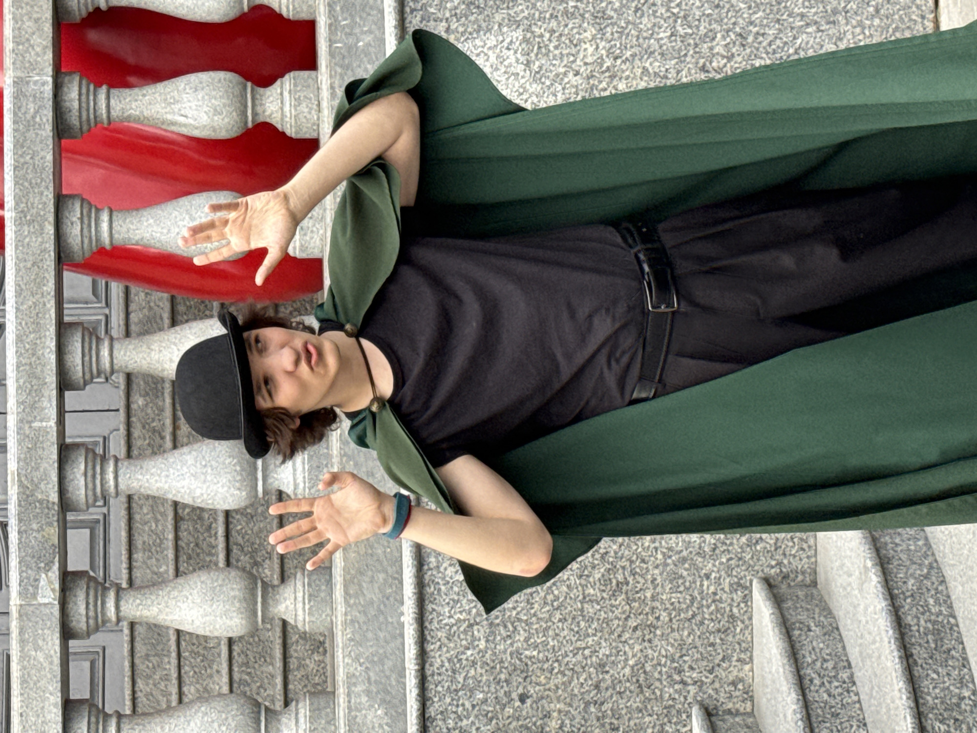 A young person, possibly a boy, is lying down on a textured surface, possibly granite or concrete, outdoors. He is wearing a black t-shirt, black pants, a black belt, a black hat, and a long, dark green cape. The cape is draped around him, covering his legs and extending over his shoulders. His right arm is bent and extended towards the camera, with his palm open and fingers spread. His left arm is also bent, with his hand in a similar open-palm gesture. He is looking towards the camera with a neutral expression.
Behind him, on the left side of the image, there is a decorative structure with vertical elements. These elements are alternating between a light gray, possibly stone, material and a vibrant red material with a wavy, layered appearance. The red sections are reminiscent of artistic representations of flames or flowing fabric. The overall scene appears to be in an urban or public space with architectural elements. The lighting suggests it is daytime, with no obvious indicators of specific weather conditions. The location context provided is Kazan, Russia. There is no visible text in the image.