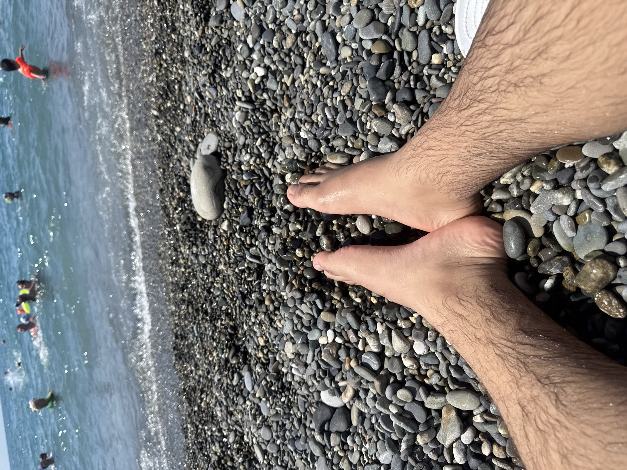The image shows a first-person perspective of someone's legs and feet resting on a pebble beach. The pebbles are of various shades of gray and are wet, indicating proximity to the water. In the background, the sea is visible with several people swimming and playing in the water. Some individuals are wearing life vests. The sea appears to be calm with gentle waves lapping at the shore. The sky is bright and appears to be sunny. The image was taken on a beach in Sochi, Russia, a resort city on the Black Sea coast.