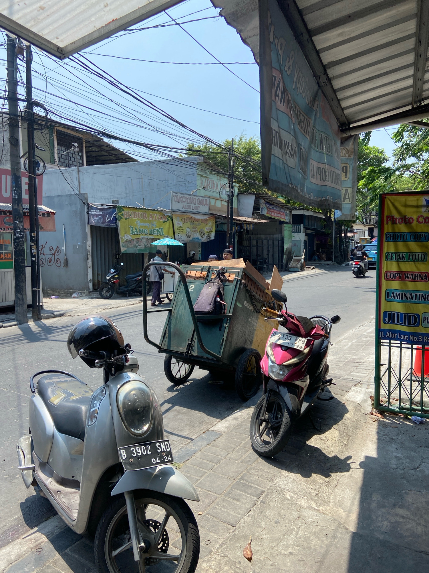 The image depicts a street scene in North Jakarta, Indonesia, under a clear, sunny sky. In the foreground, a silver scooter with a black helmet on its handlebars is parked on a cobblestone sidewalk. To the right of the scooter, a pink scooter is parked. Behind the scooters, a large, green, two-wheeled cart filled with various items, possibly including cardboard and fabric, is positioned on the street. A man in a light-colored shirt and dark pants is walking behind the cart, pushing it.
The street is lined with buildings, some of which appear to be shops or businesses. A banner hangs from a metal awning, displaying text in Indonesian that is difficult to read due to the angle and lighting. Another vertical banner on the right side of the image advertises "Photo Copy," "Cetk Foto," "Copy Warna," "Laminating," and "Jilid." Further down the street, several other motorbikes and a light blue car are visible. Overhead, a dense network of electrical wires crisscrosses the sky, a common sight in urban Indonesian environments. The overall environment suggests a bustling commercial area.