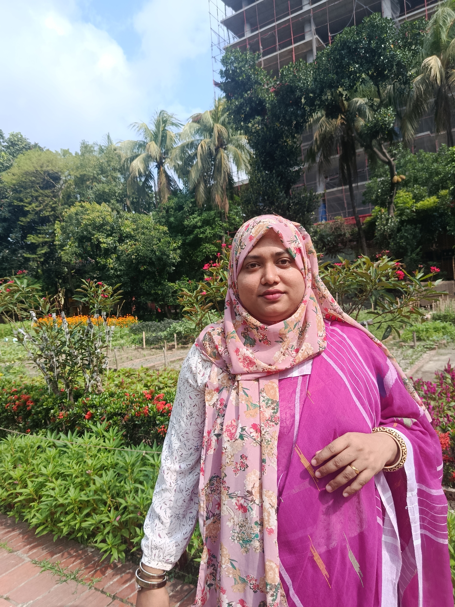 A middle-aged woman is standing in a garden, possibly in Dhaka, Bangladesh. She is wearing a pink floral hijab and a pink and white sari with long sleeves. She has gold bangles on her left wrist and a gold ring on her right hand. Behind her is a garden with various plants and flowers. A partially constructed building is visible in the background, surrounded by trees including palm trees. The sky is visible with some clouds. A brick path is visible in the foreground.