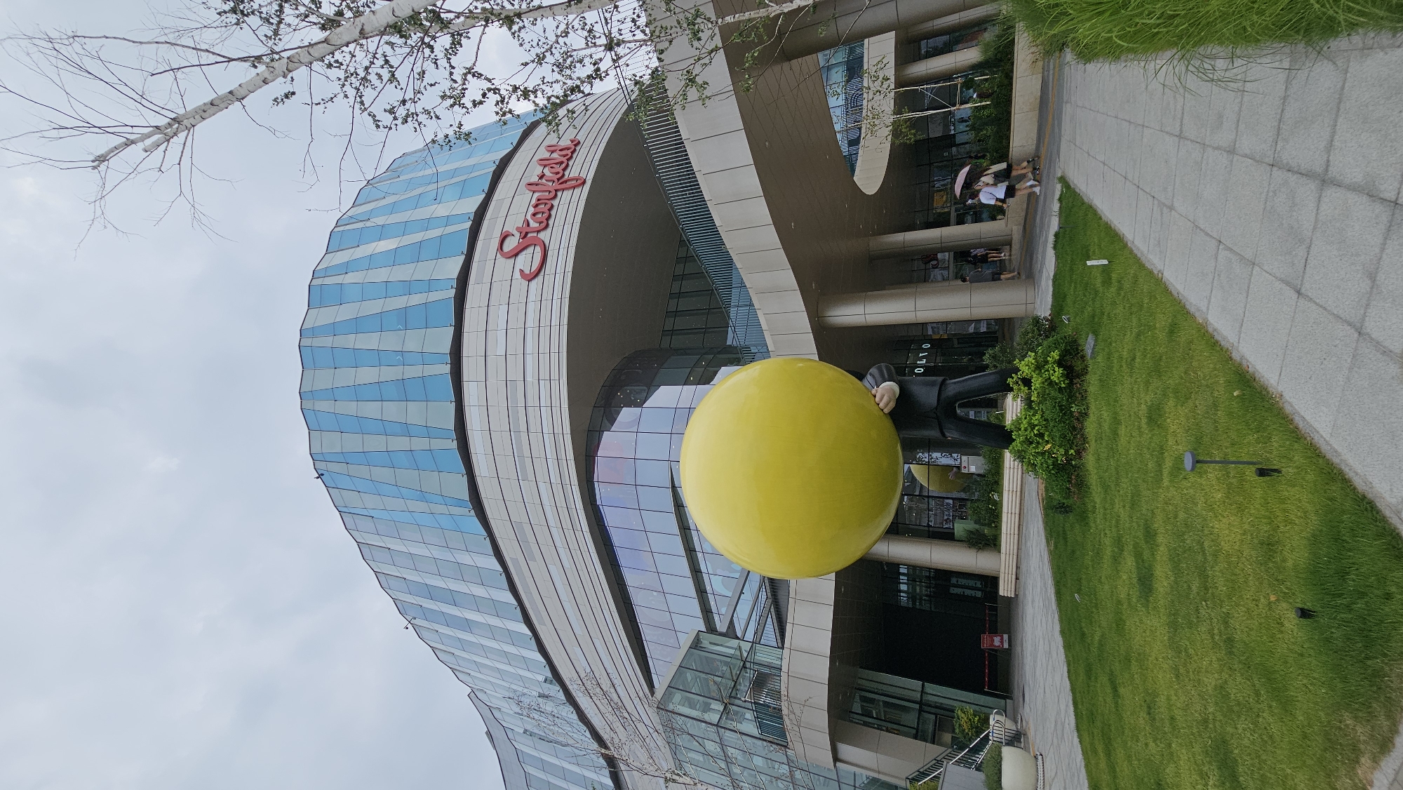 An external low-angle shot captures the Starfield Hanam mall in Hanam-si, Gyeonggi-do, South Korea. The mall's facade features a prominent, curving design with panels of alternating blue glass and white material. Above the entrance, the "Starfield" logo is displayed in red lettering. In the foreground, a large, bright yellow sphere is held aloft by a statue of a person in a black suit. The statue stands on a patch of green grass, bordered by a paved walkway. Several trees with bare branches are visible to the right of the structure. The sky is overcast. In the background, a few individuals, including one holding a pink umbrella, walk along the pathway.