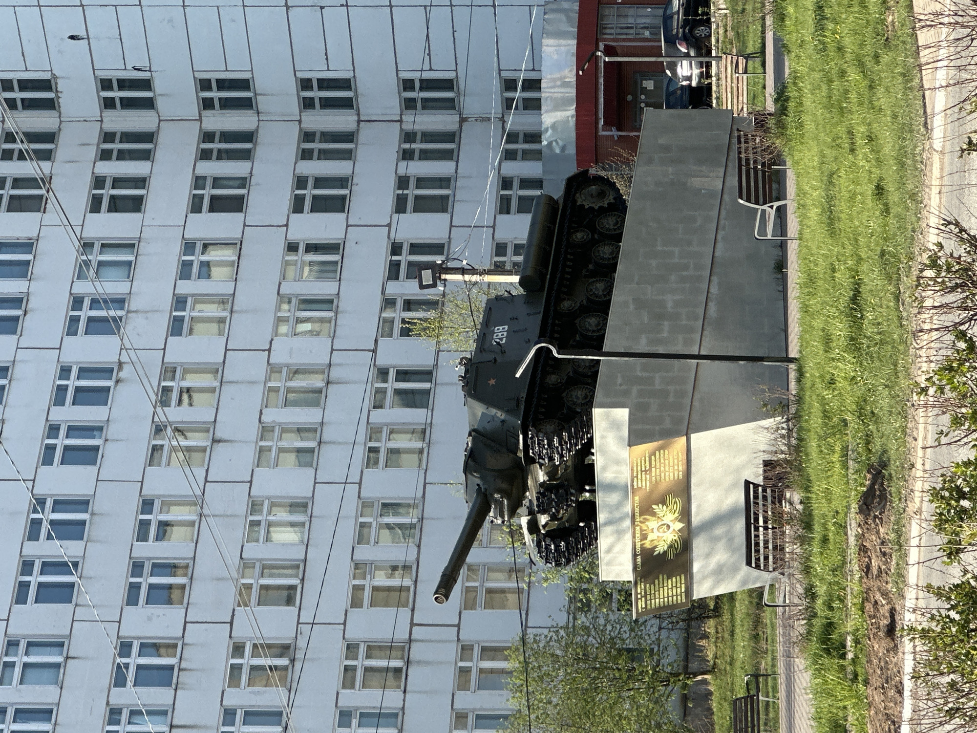 The image captures a Soviet IS-2 tank monument positioned in front of a large, grey apartment building in Chaikovsky, Russia. The tank, painted in dark green, is mounted on a concrete platform. A plaque bearing text and an emblem adorns the monument base. The surrounding environment includes grass, trees, and a paved walkway with benches, suggesting a park or memorial area. A nearby building with a red façade is also visible. The lighting suggests it is daytime.