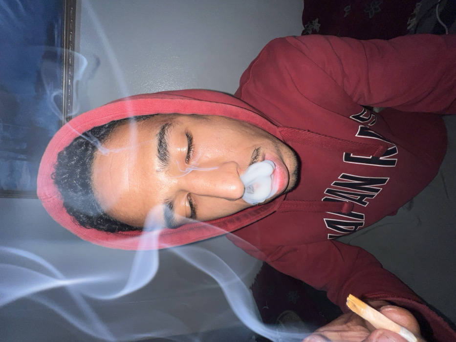 A male individual with closed eyes, wearing a dark red hooded sweatshirt, is captured mid-exhale, producing a prominent smoke ring from his mouth. Wisps of smoke are visible around his face and head. His left hand holds a hand-rolled cigarette (joint) with a visible filter. The background indicates an indoor residential setting, featuring a light-colored wall, a framed picture with a muted blue-grey scene, and portions of a dark red patterned rug or textile. This scene takes place within a private residence in Tanger, Morocco.
