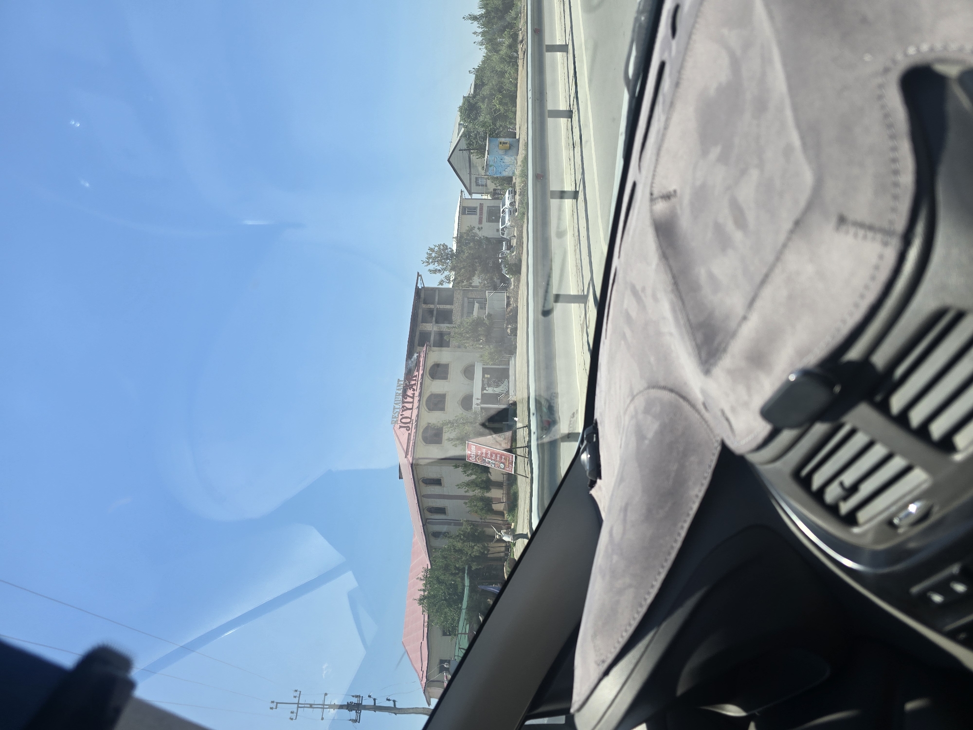 The image is taken from inside a vehicle, looking out through the windshield. The view encompasses a roadside scene with a large, light-colored building with a red roof that has signage reading "RESTAURANT JOYIZAN". A person is standing near the building, and a red A-frame sign is positioned in front of it, displaying text in a non-English script, likely Uzbek. Beyond the building, there are trees and other structures, including what appears to be a smaller building with a yellow utility box in front of it, and cars parked along the road. The road itself is visible with a metal guardrail separating it from the area with the buildings. The sky is clear and blue, indicating a sunny day. The foreground shows the interior of the car, including the dashboard with a grey, textured cover and air vents. The location is identified as Safkarda, Uzbekistan.