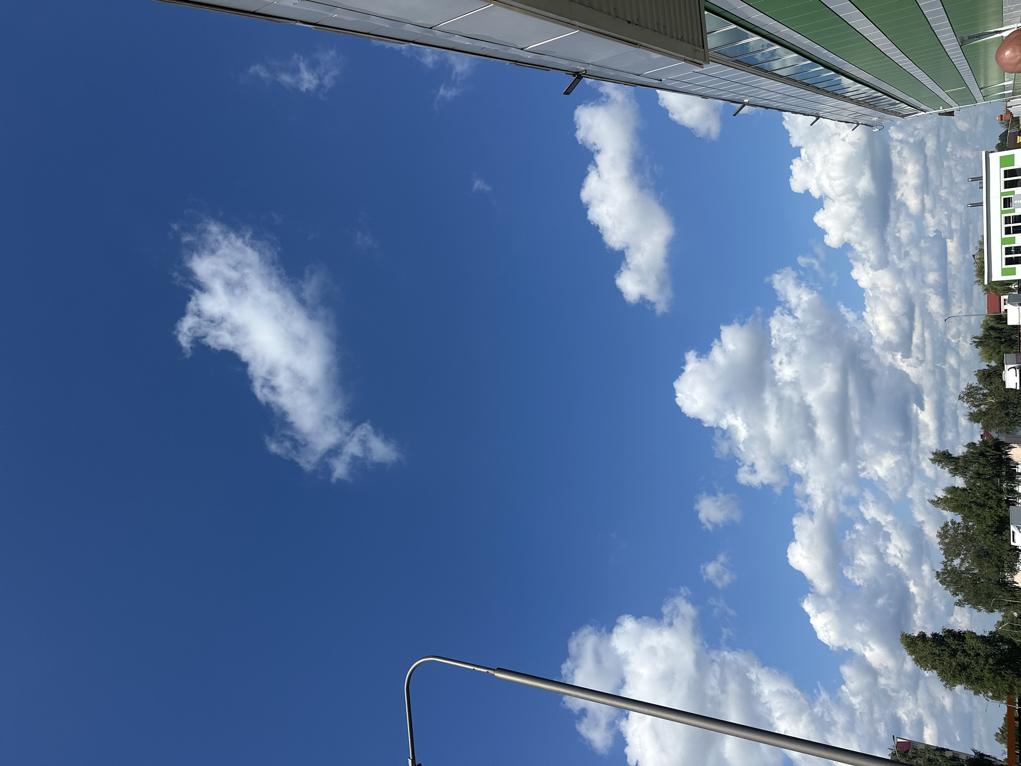 The image captures a bright, clear blue sky with scattered white cumulus clouds. A tall, gray lamppost is positioned on the left side of the frame, extending upwards and curving out of view. To the right, the side of a modern building is visible, featuring a green facade with horizontal white stripes and a row of windows reflecting the sky. Below the sky and the building, a line of green trees is visible, interspersed with some buildings and what appear to be vehicles. The scene suggests an outdoor urban or suburban environment on a sunny day. Based on the context provided, this scene is located in the city of Maladzyechna, Belarus. No people are directly visible in the frame.