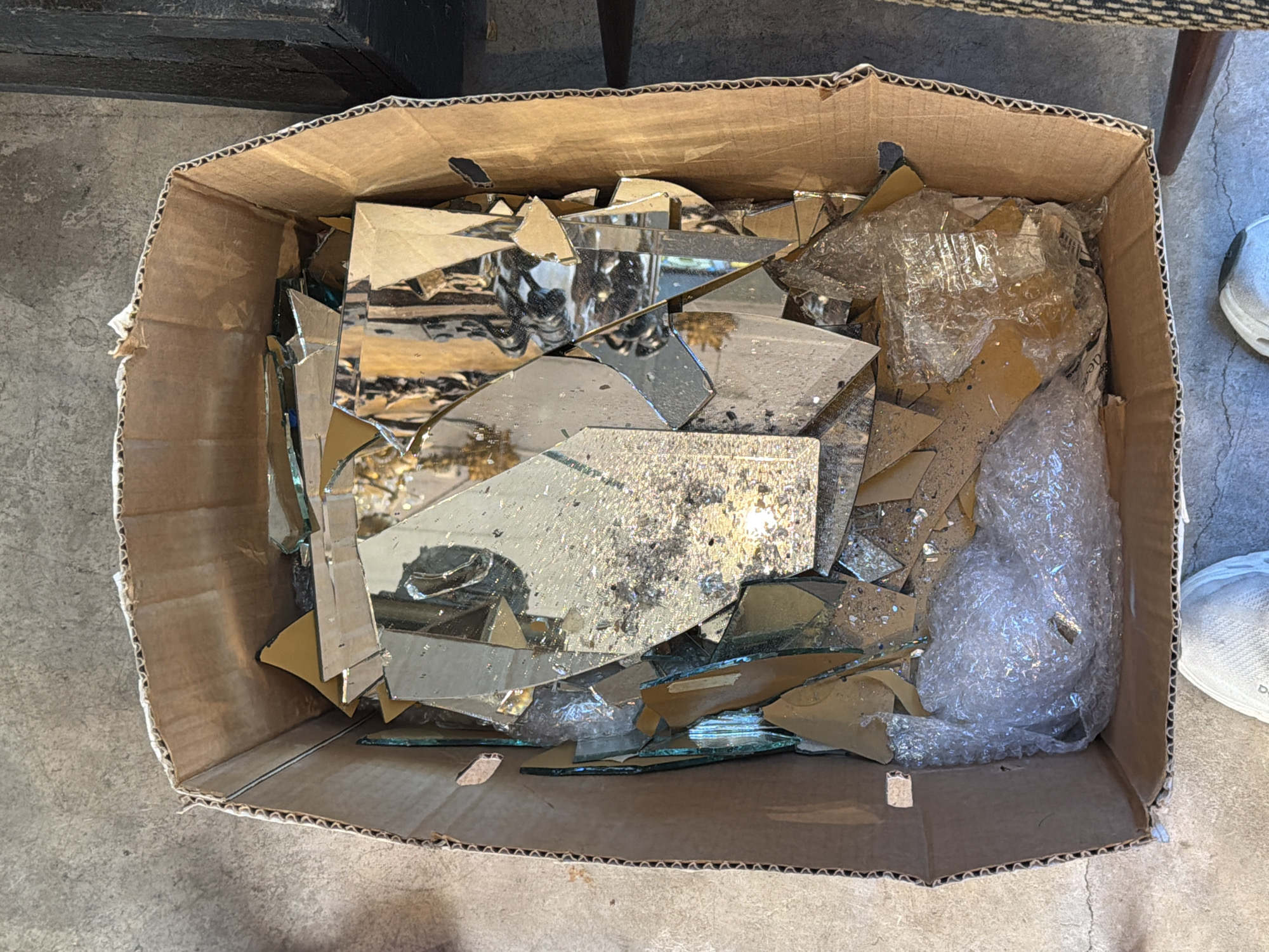 A cardboard box is filled with broken pieces of a reflective material, likely glass or mirror, with some pieces wrapped in bubble wrap. The box sits on a concrete surface. A leg of a chair can be seen in the background, and a sneaker is partially visible on the right side. The location is unspecified and there's no clear evidence pointing to the city of Buenos Aires, Argentina.