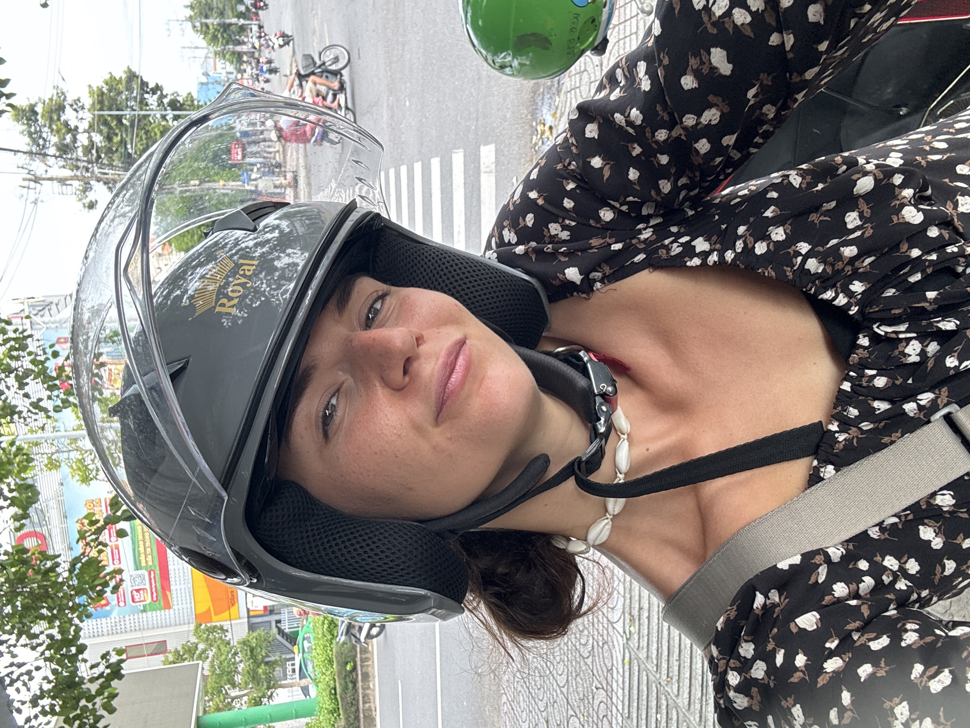 The image presents a close-up portrait of a young woman situated in an urban environment, likely in Phu Thuy Ward, Vietnam. She looks directly at the viewer with a neutral expression. She is wearing a black or dark grey open-face motorcycle helmet with a clear visor, which features a gold "Royal" logo topped with a crown on its side. Around her neck, she has a necklace made of white shells. Her attire consists of a long-sleeved black top or dress patterned with white and brown flowers. A grey strap, possibly from a bag or a vehicle harness, crosses her chest.
The background reveals a bustling street scene, characteristic of an urban area. A white pedestrian crosswalk is visible on the asphalt road. In the distance, several motorbikes and a bicycle lying on its side are scattered. Trees, buildings, and indistinct commercial signs line the street. A bright green object, possibly another helmet or part of a motorbike, is partially visible near her right shoulder. The daylight illumination suggests the photo was taken during the day. The overall impression is that she is a passenger on a motorbike, perhaps stopped in traffic or waiting by the roadside.