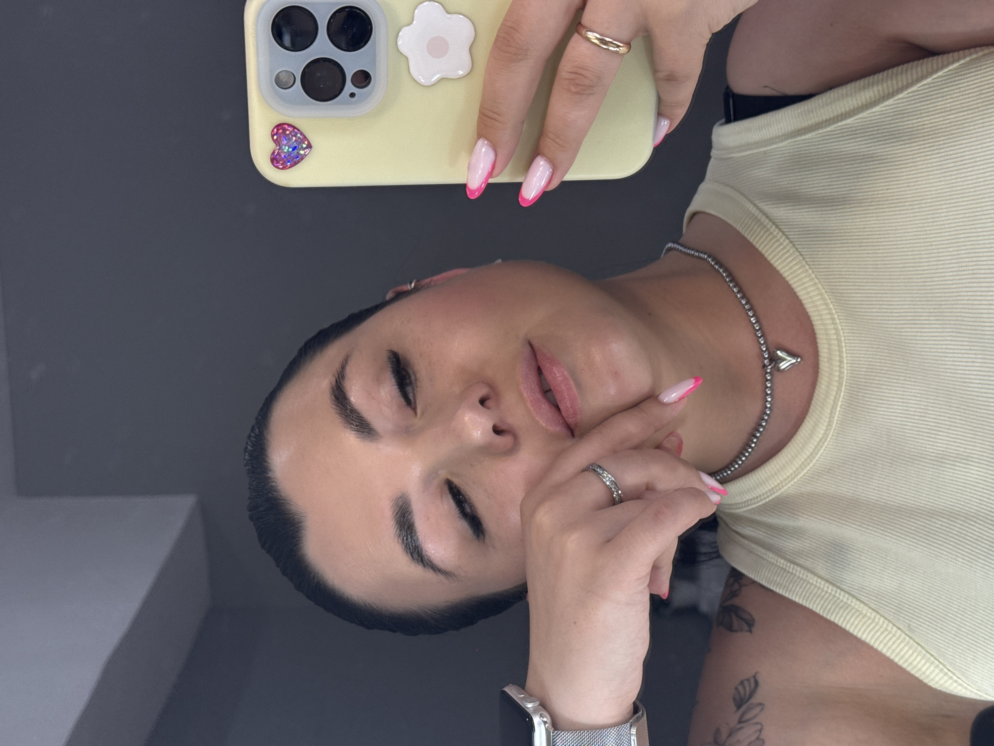 A selfie depicts a light-skinned woman with dark hair pulled back, wearing a light yellow ribbed tank top and a silver necklace. She holds a yellow phone with a heart sticker and a daisy accessory in her hand, showcasing pink-tipped nails. She also wears a silver watch and a ring on her finger. The environment is dark, possibly a room interior. There is no information to deduce the location to be Chaikovsky, Russia.