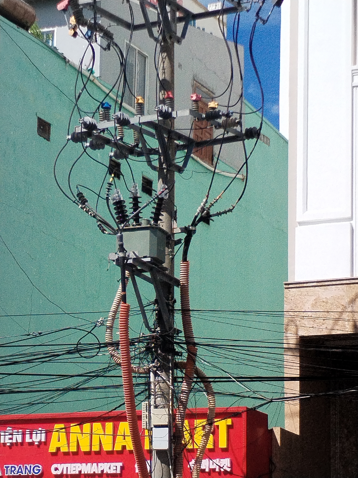 This photograph captures a busy urban scene, dominated by a central utility pole laden with a dense network of electrical wires and a transformer. The pole, made of concrete, supports multiple cross arms from which numerous black wires extend in all directions, creating a chaotic web. Various insulators, some clear, some black, and others with distinct colored caps (red, yellow, blue), are visible at different points on the pole, managing the connections. A gray electrical transformer is mounted on the lower-mid section of the pole, with thick, corrugated orange and white conduits running vertically alongside it, protecting some of the main cables.
In the background, two buildings are visible. To the left, a building with a textured, teal-green painted wall spans much of the frame. Above it, part of a lighter-colored building with windows can be seen. To the right, a white building with prominent vertical paneling stands, contrasting with a lower section of beige or brown concrete. The bright sunlight suggests it is daytime, casting clear shadows.
At the bottom of the image, a large red sign with yellow block letters is partially visible. The text clearly reads "TIỆN LỢI ANNA" (Convenient ANNA), with "NHA TRANG" and "СУПЕРМАРКЕТ" (SUPERMARKET) in Cyrillic script below it, indicating a commercial establishment. The overall scene reflects a common urban landscape in areas like Nha Trang, Vietnam, characterized by a complex overhead utility infrastructure.